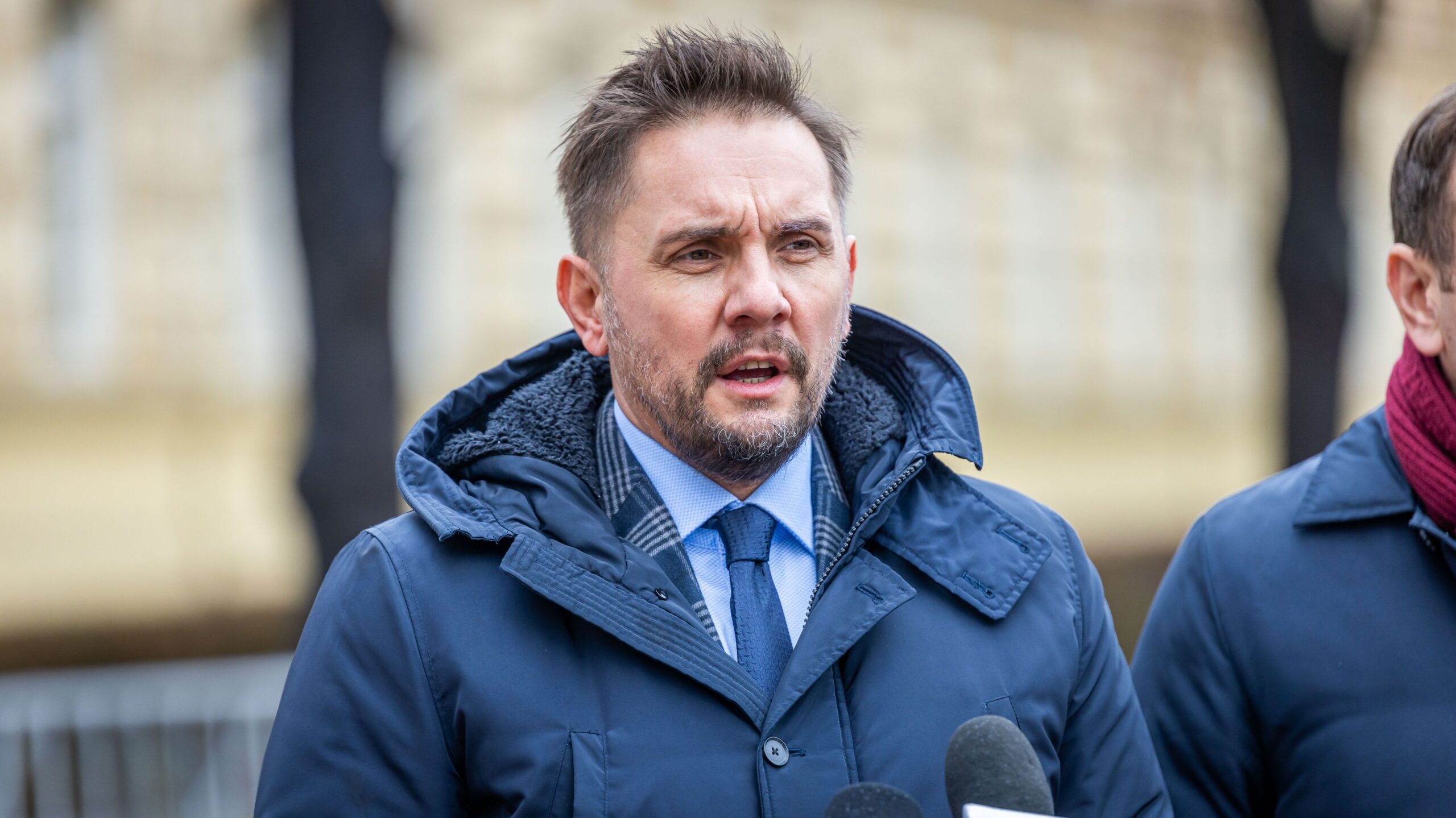 Mężczyzna w granatowej kurtce z kapturem wygłasza oświadczenie przed mikrofonami.