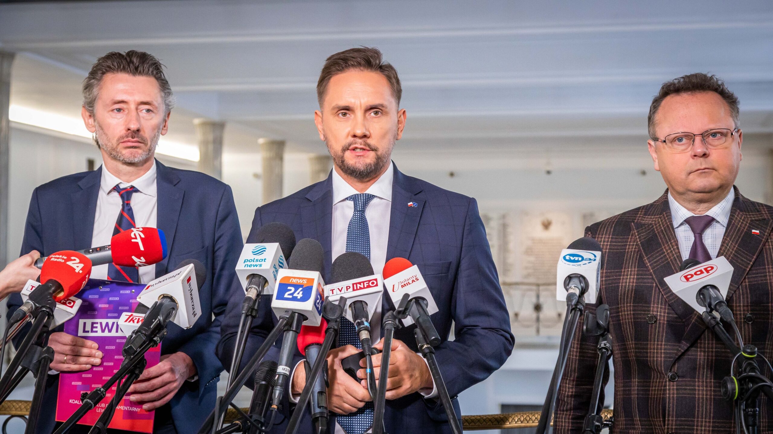 Trzech polityków stoi przed mikrofonami podczas konferencji prasowej w formalnym otoczeniu.