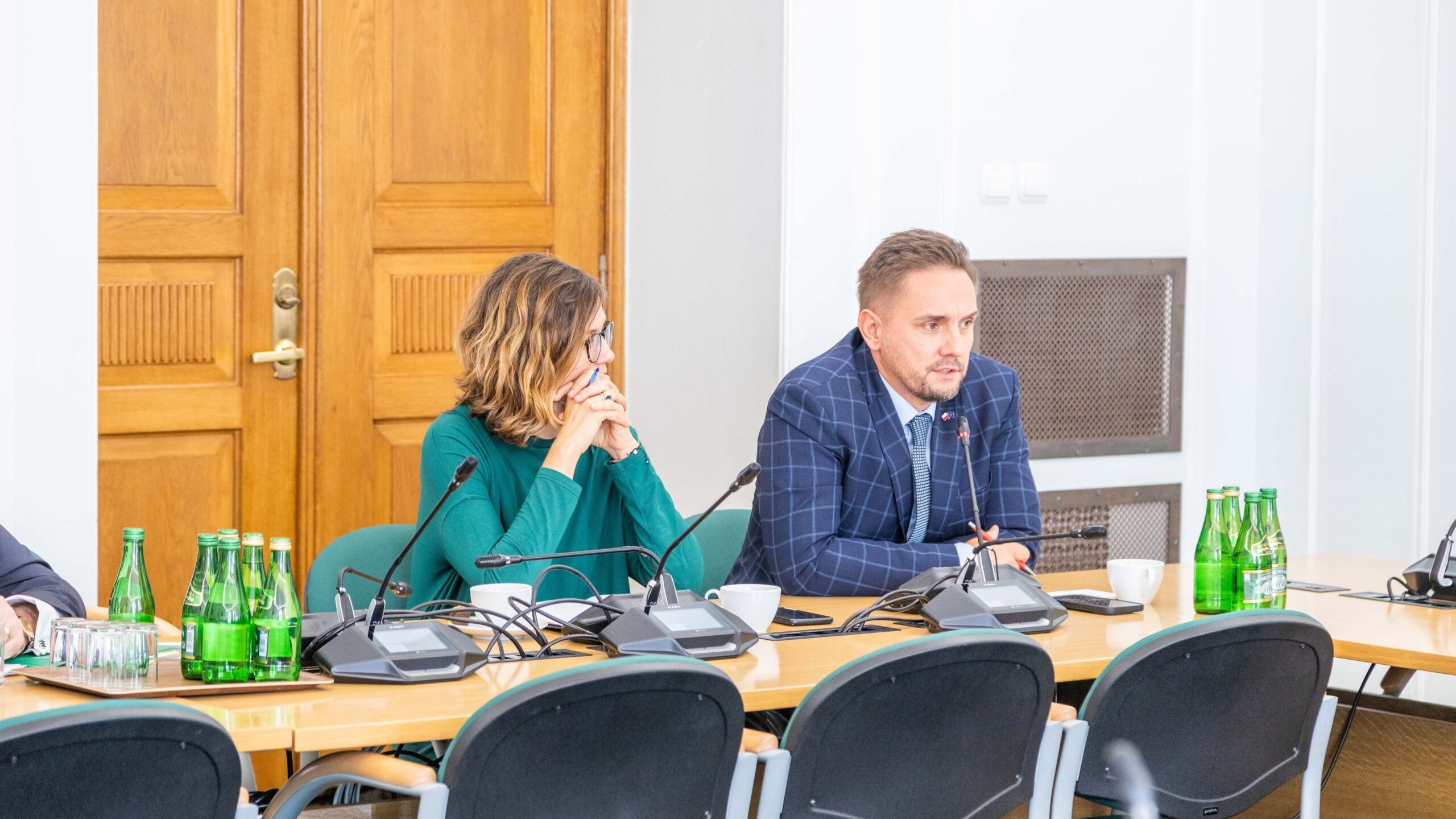 Na zdjęciu widać dwóch ludzi siedzących przy stole konferencyjnym, z butelkami wody i mikrofonami na stole, a w tle widoczne są drewniane drzwi.