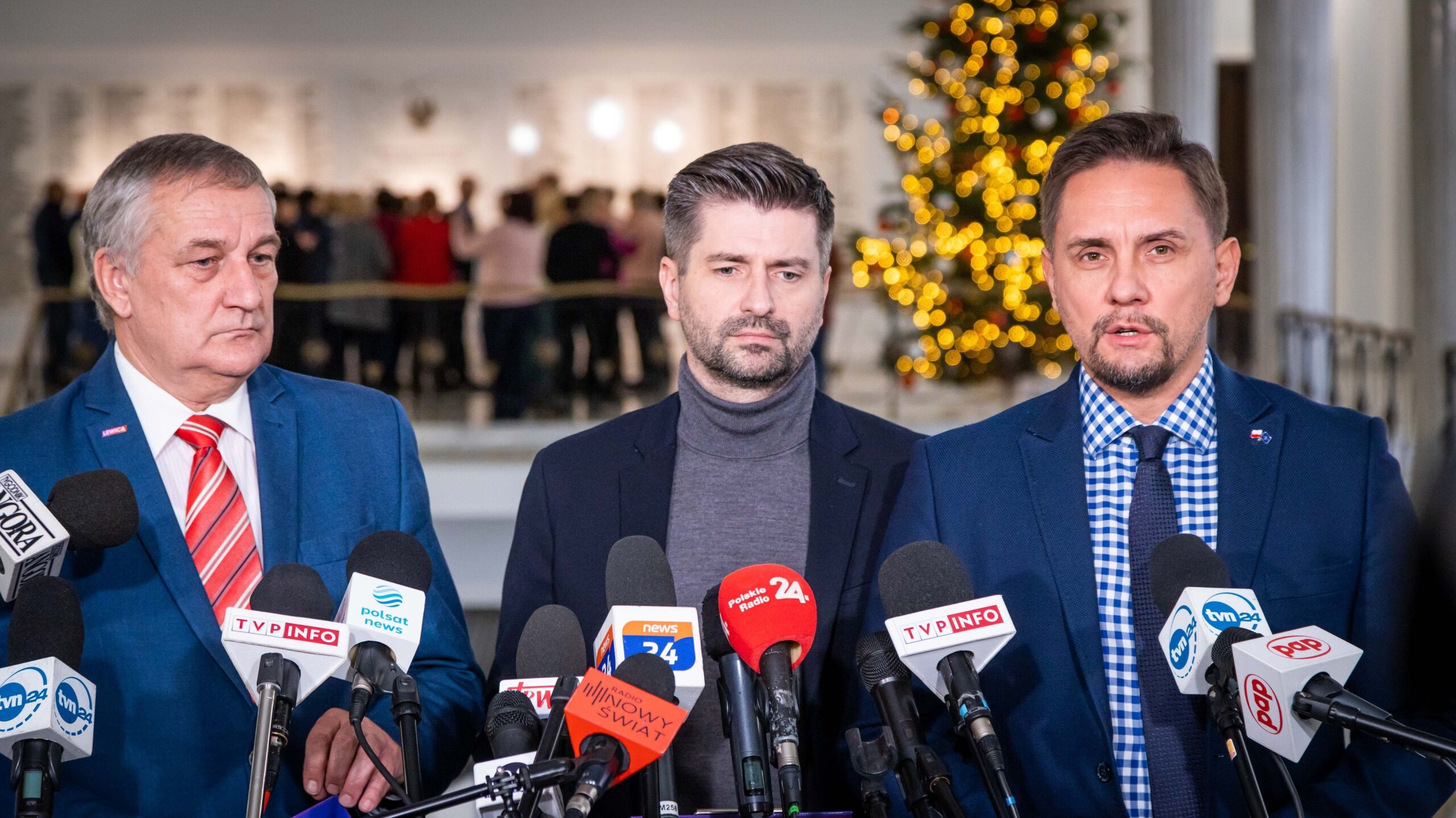 Trzech mężczyzn w garniturach udziela konferencji prasowej, stojąc przed licznymi mikrofonami; w tle widoczna dekorowana choinka i grupa osób.