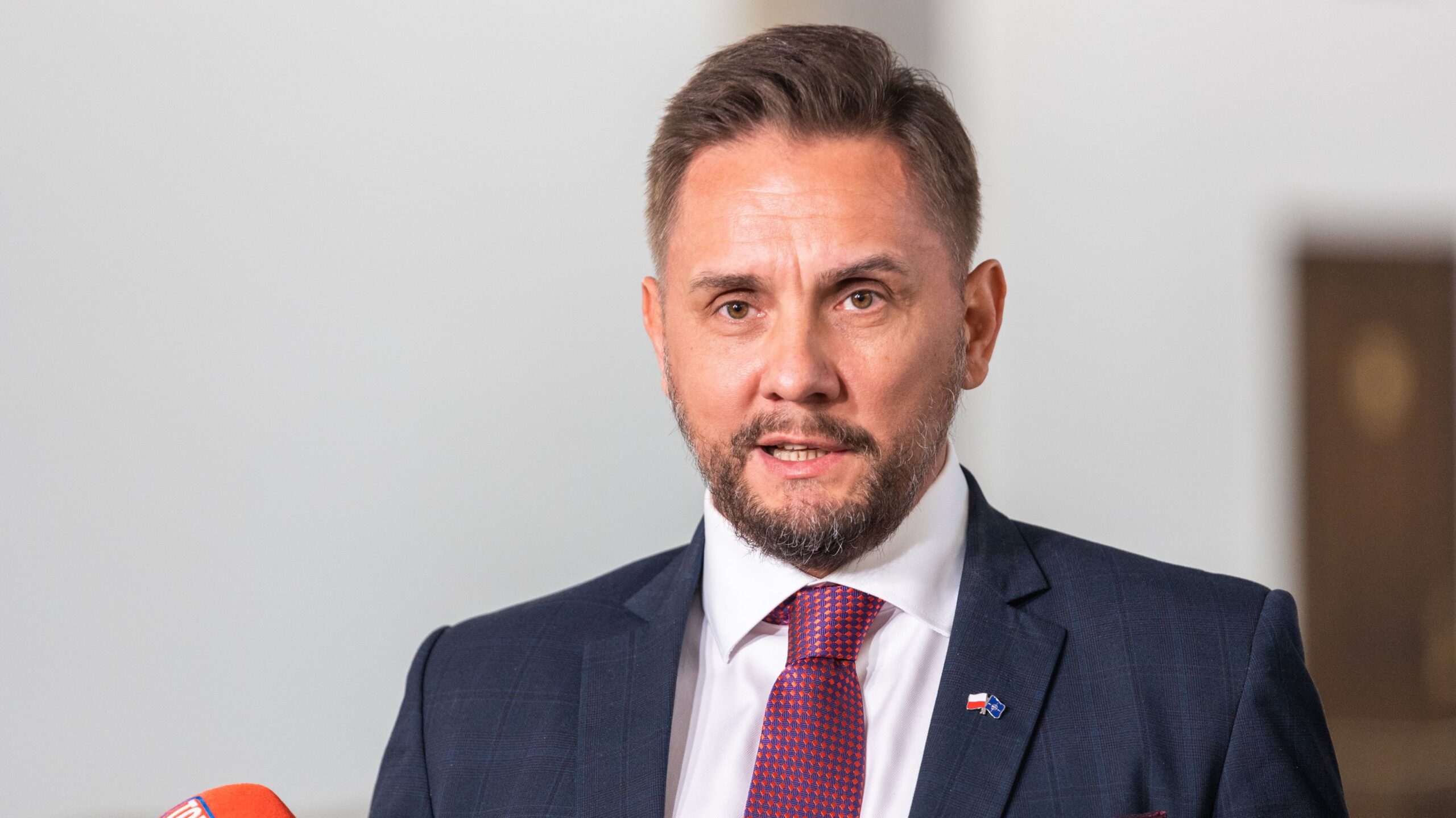 Mężczyzna w garniturze z brodą mówi do kamery, trzymając mikrofon w tle jasnego pomieszczenia.