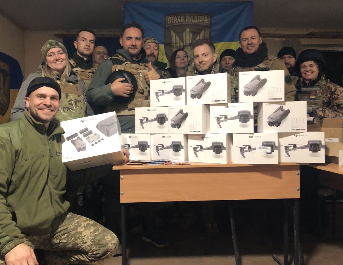Na zdjęciu widać grupę osób w mundurach, które pozują z wieloma pudełkami z dronami w tle, a za nimi powiewa flagą Ukrainy.