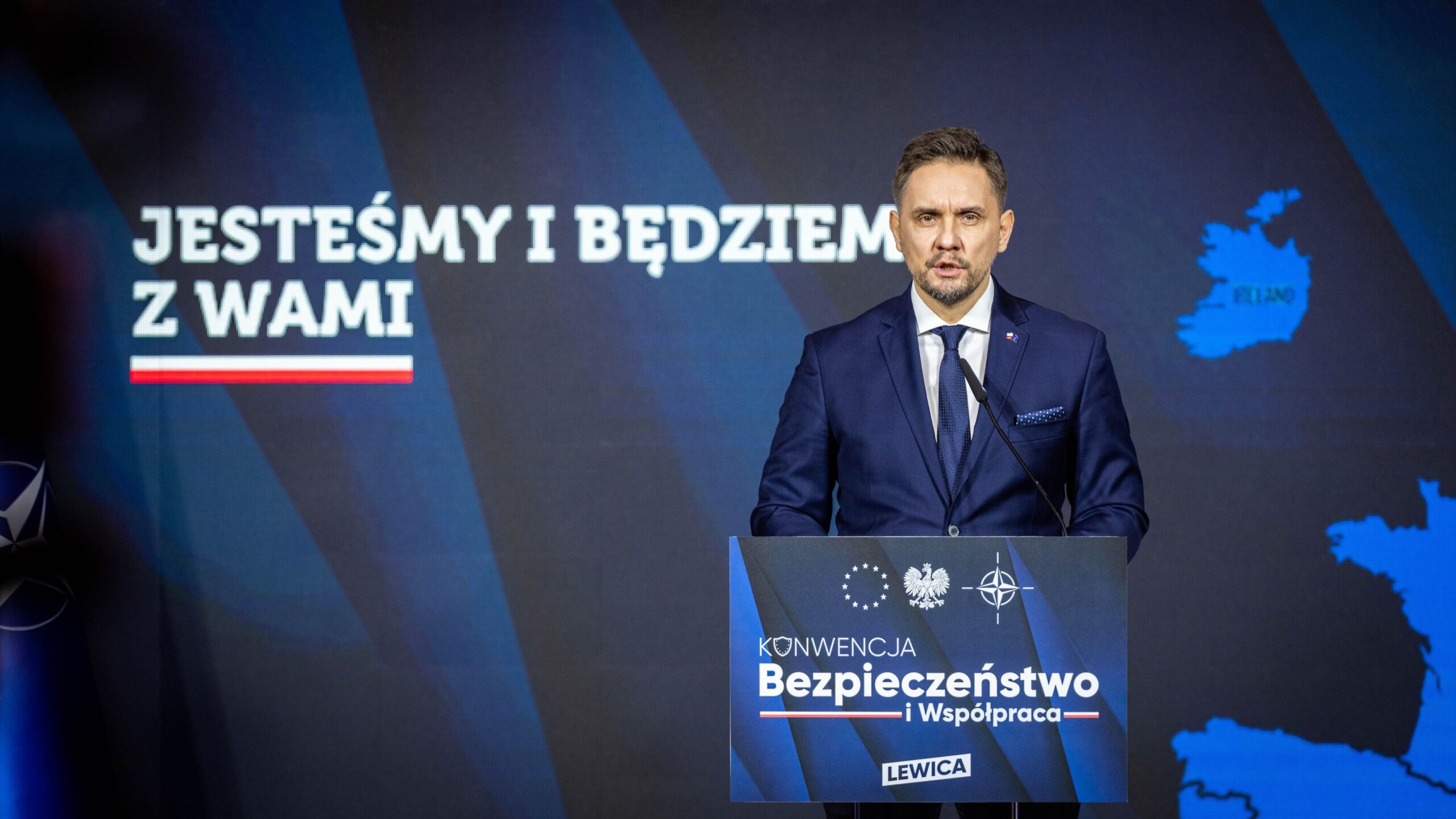Mężczyzna w granatowym garniturze przemawia z podium podczas konferencji o bezpieczeństwie Polski, na tle baneru z napisem „Bezpieczeństwo i Współpraca” i mapą Polski.