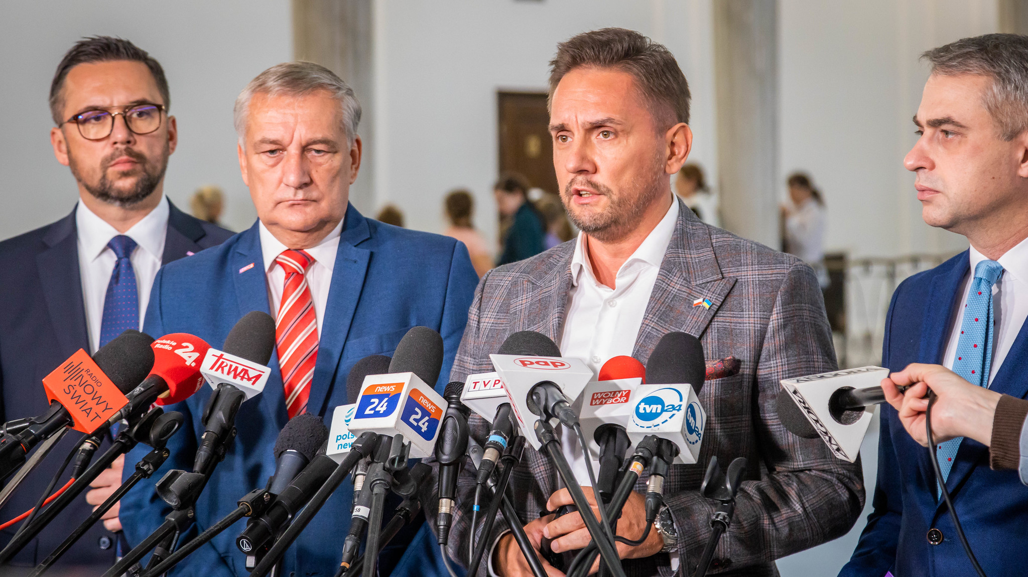 Na zdjęciu znajdują się czterech mężczyzn ubranych w garnitury, stojących przed mikrofonami podczas konferencji prasowej.