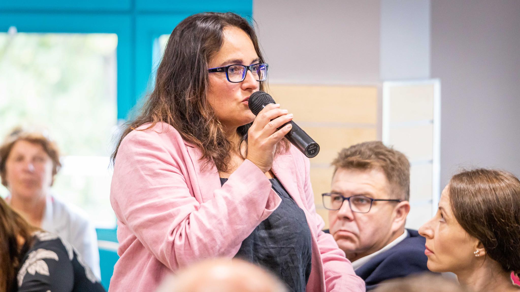 Kobieta w różowej marynarce i okularach mówi do mikrofonu podczas spotkania o bezpieczeństwie mieszkańców wzdłuż Odry.