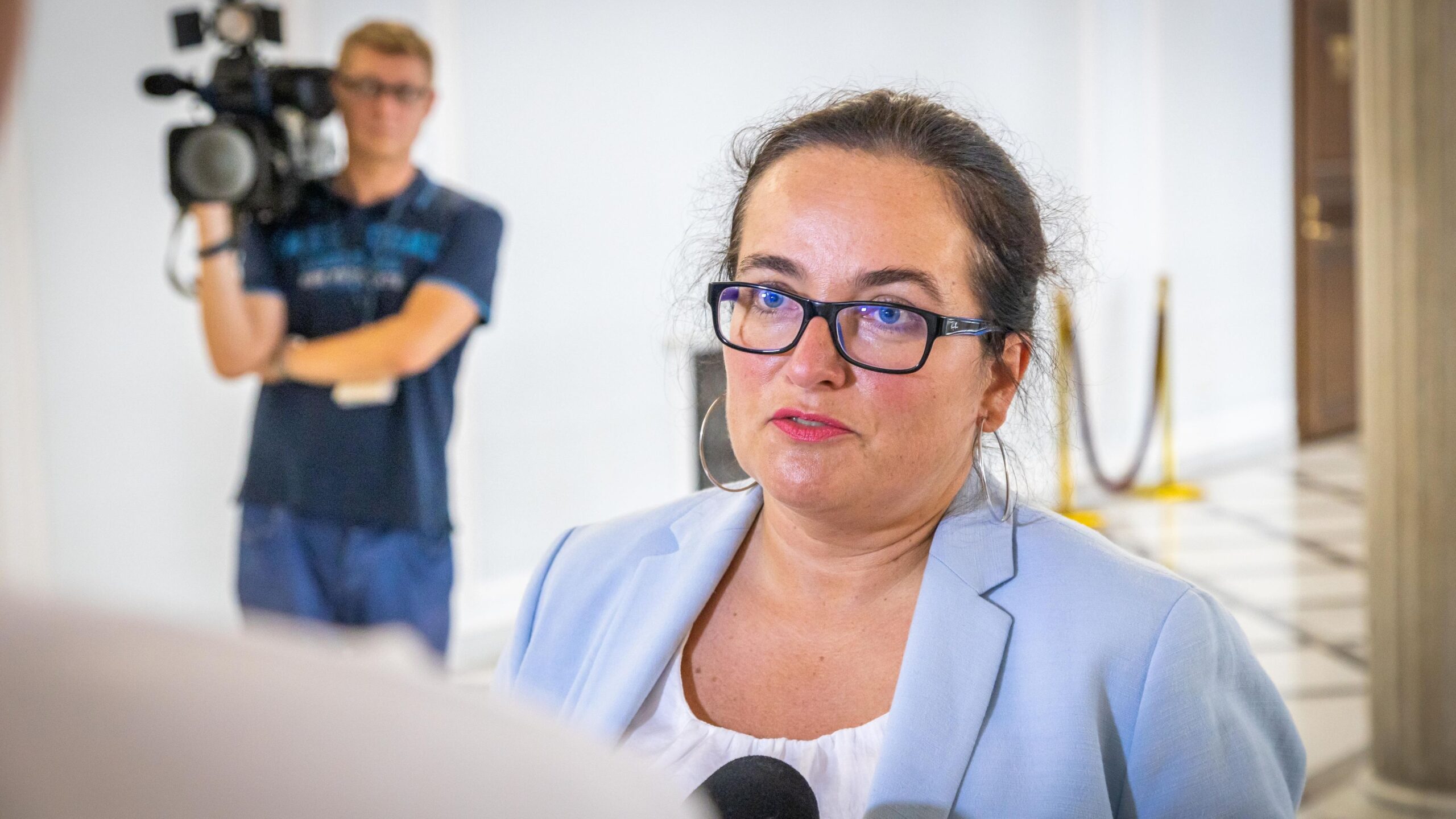 Na zdjęciu widoczna jest kobieta z okularami, ubrana w jasną marynarkę, która udziela wywiadu, podczas gdy w tle stoi operator kamery.