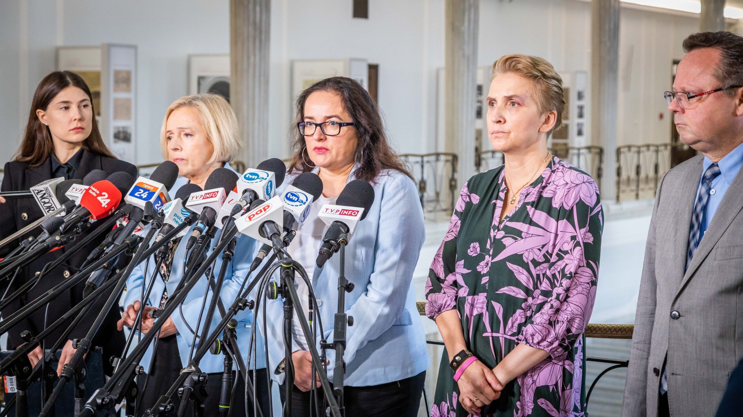 Na zdjęciu widać grupę osób występujących na konferencji prasowej, z mikrofonami w tle i różnorodnymi wyrazami twarzy.