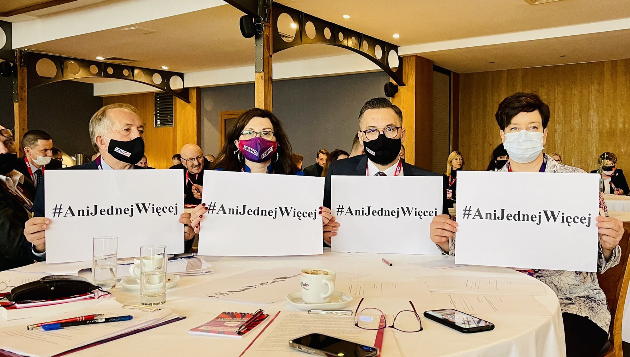 Grupa uczestników konferencji siedzących przy stole, trzyma kartki z hashtagiem #AniJednejWięcej i nosi maseczki.