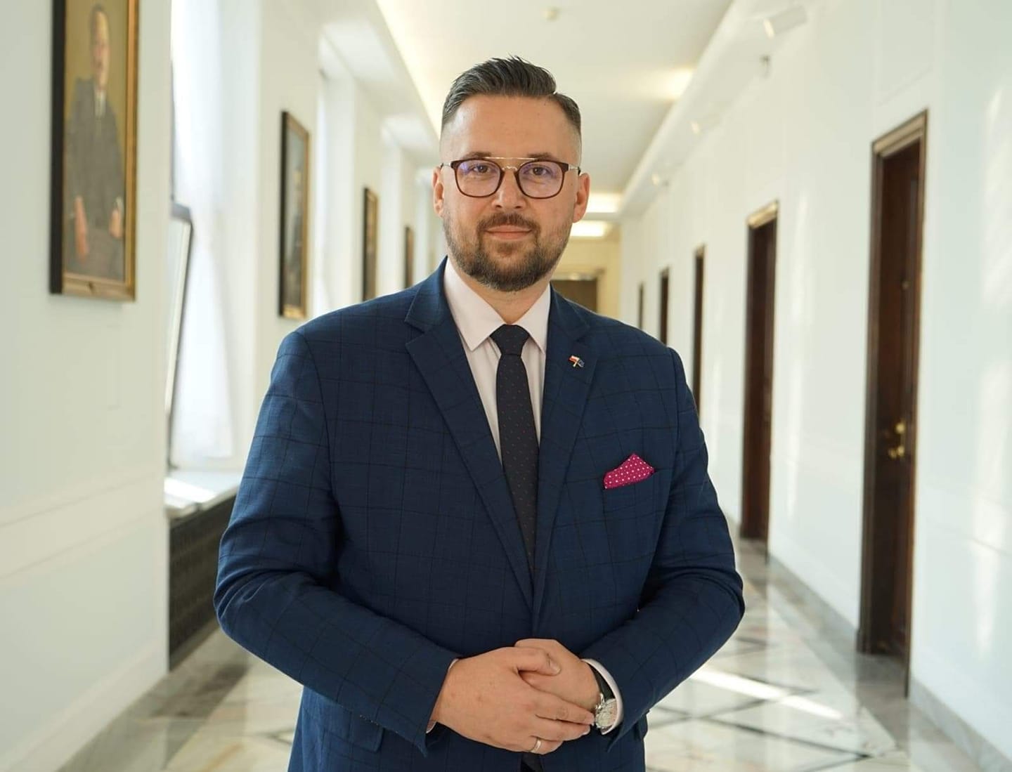 Mężczyzna w niebieskim garniturze z krawatem i kieszonkową chusteczką stoi w eleganckim korytarzu z obrazami na ścianach.