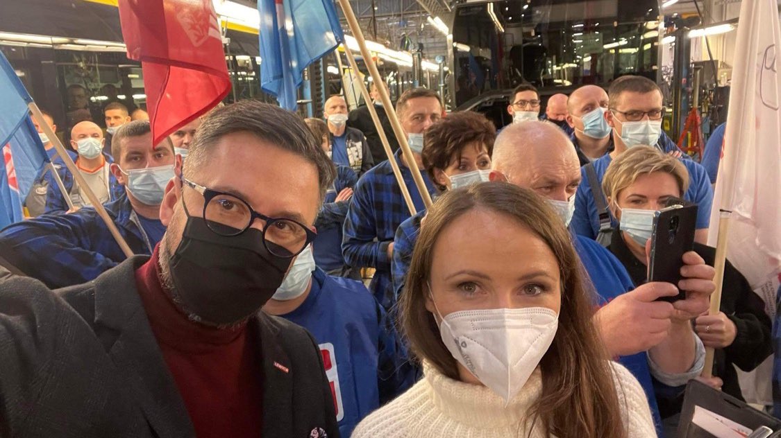 Grupa ludzi nosi maski ochronne i trzyma flagi podczas wydarzenia, na pierwszym planie dwie osoby pozują do selfie.