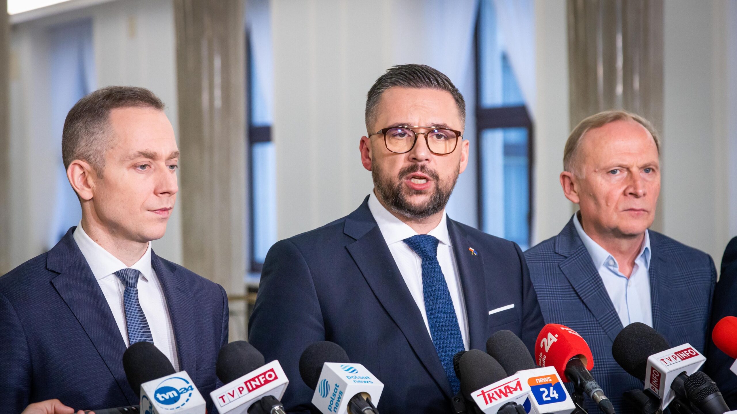 Trzech mężczyzn w garniturach stoi przed mikrofonami podczas konferencji prasowej.