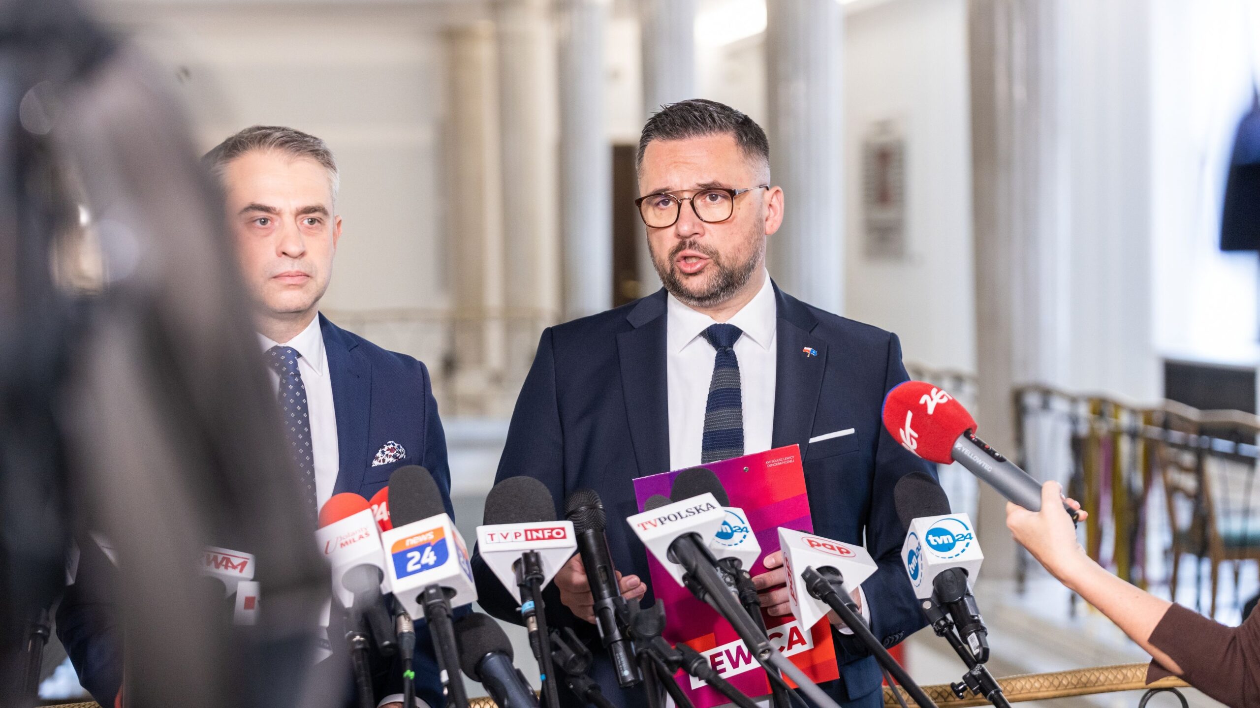 Dwóch mężczyzn w garniturach udziela konferencji prasowej, stojąc za licznymi mikrofonami mediów w jasnym holu z kolumnami.