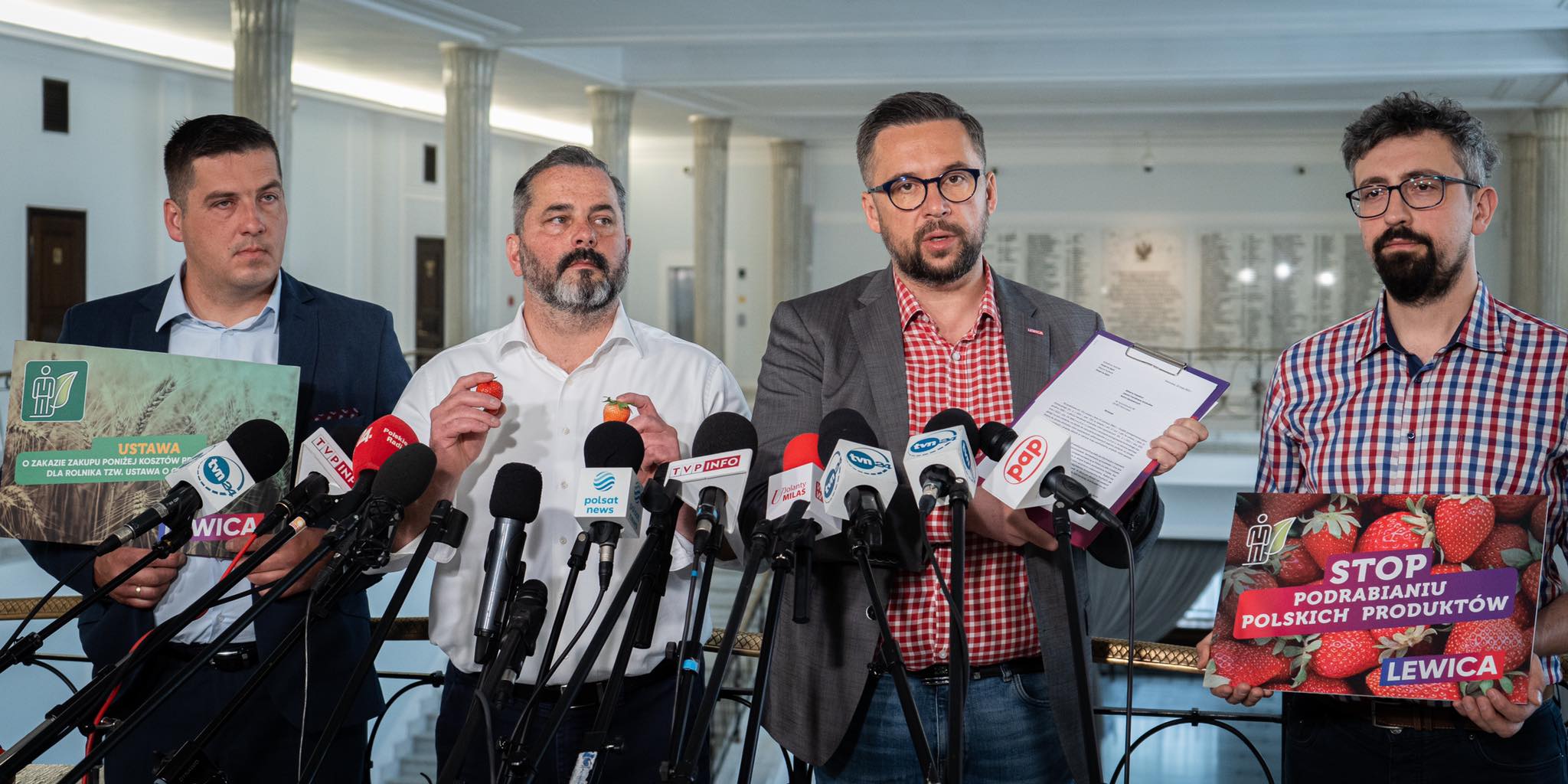 Na zdjęciu czterech mężczyzn w garniturach, stojących za mikrofonami, trzymających plakaty związane z ustawą dotyczącą ochrony polskich produktów rolnych oraz ich promowaniem.