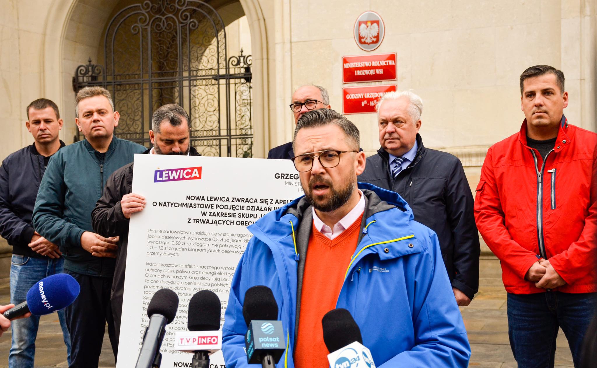 Grupa mężczyzn podczas konferencji prasowej przed wejściem do urzędu, na pierwszym planie mówca w niebieskiej kurtce i pomarańczowym swetrze mówiący do mikrofonów, za nim plakat z logiem Lewicy dotyczący interwencyjnego skupu jabłek.