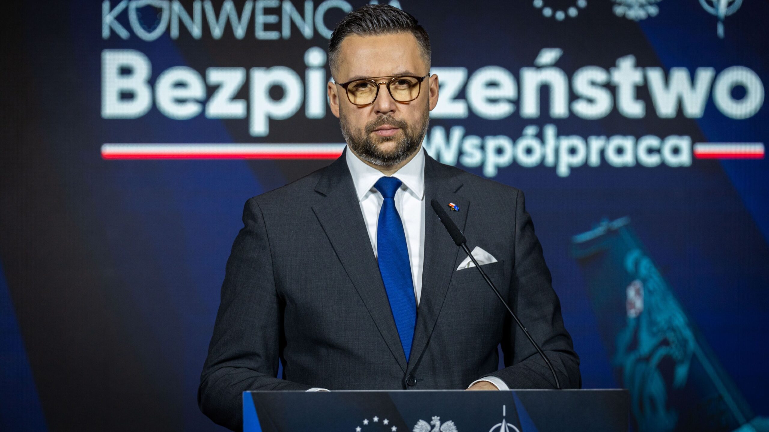 Mężczyzna w garniturze przemawia z mikrofonem z mównicy podczas konferencji o bezpieczeństwie, na tle dużych napisów „Bezpieczeństwo” i „Współpraca”.