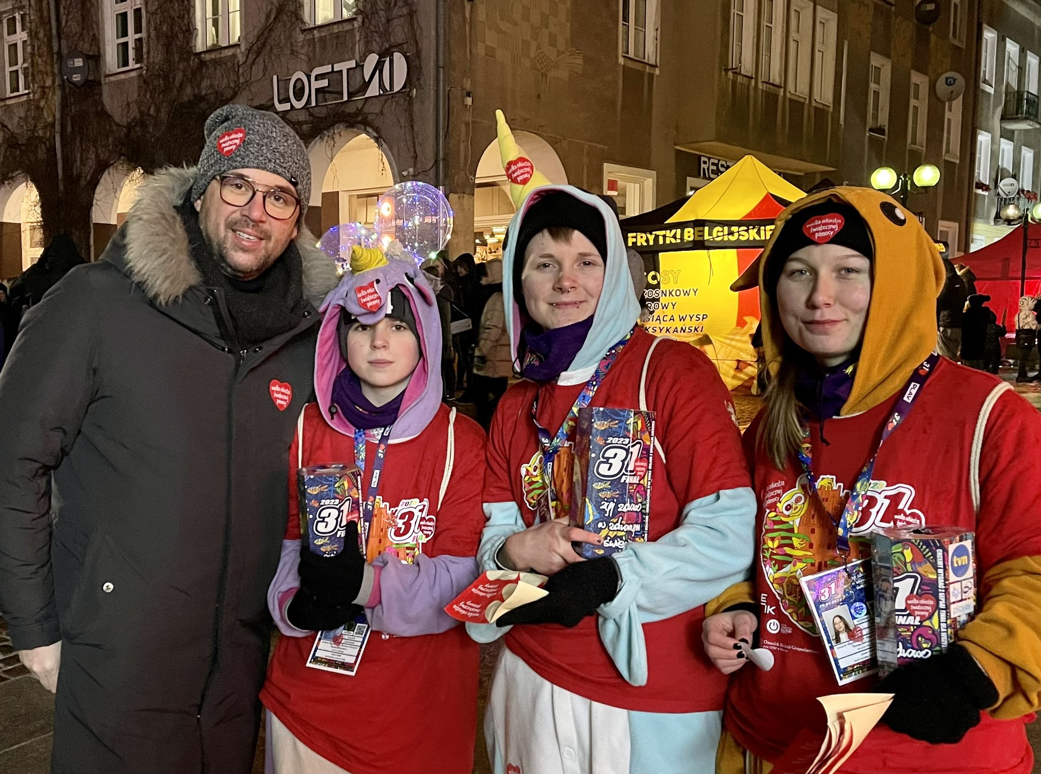 Czterech wolontariuszy WOŚP w czerwonych koszulkach z identyfikatorami i przebraniach stoi na miejskiej ulicy podczas finału WOŚP 2023, trzymając kolorowe pudełka z numerem 31.