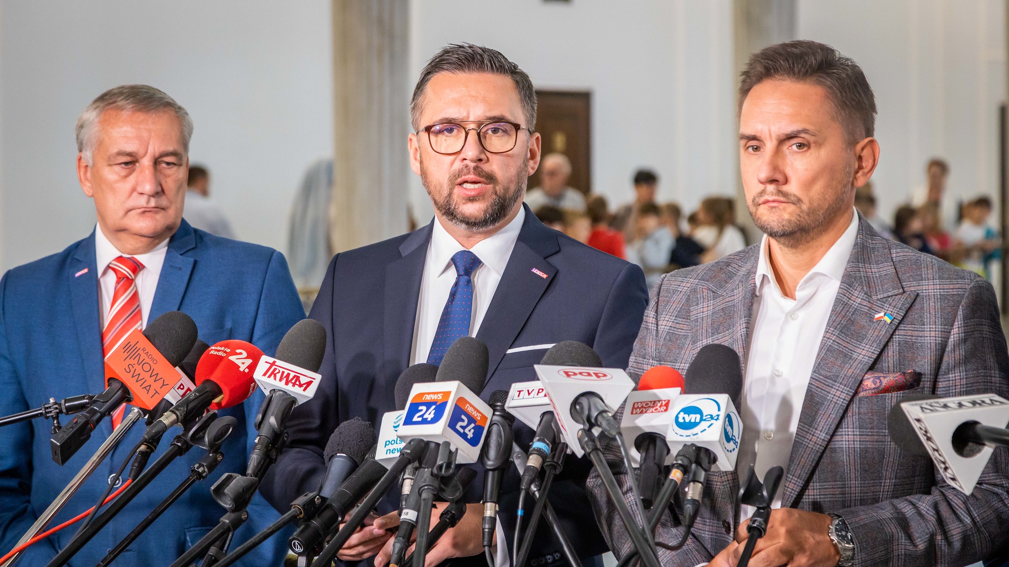 Na zdjęciu trzej mężczyźni stoją przed mikrofonami podczas konferencji prasowej, w tle widać tłum ludzi.