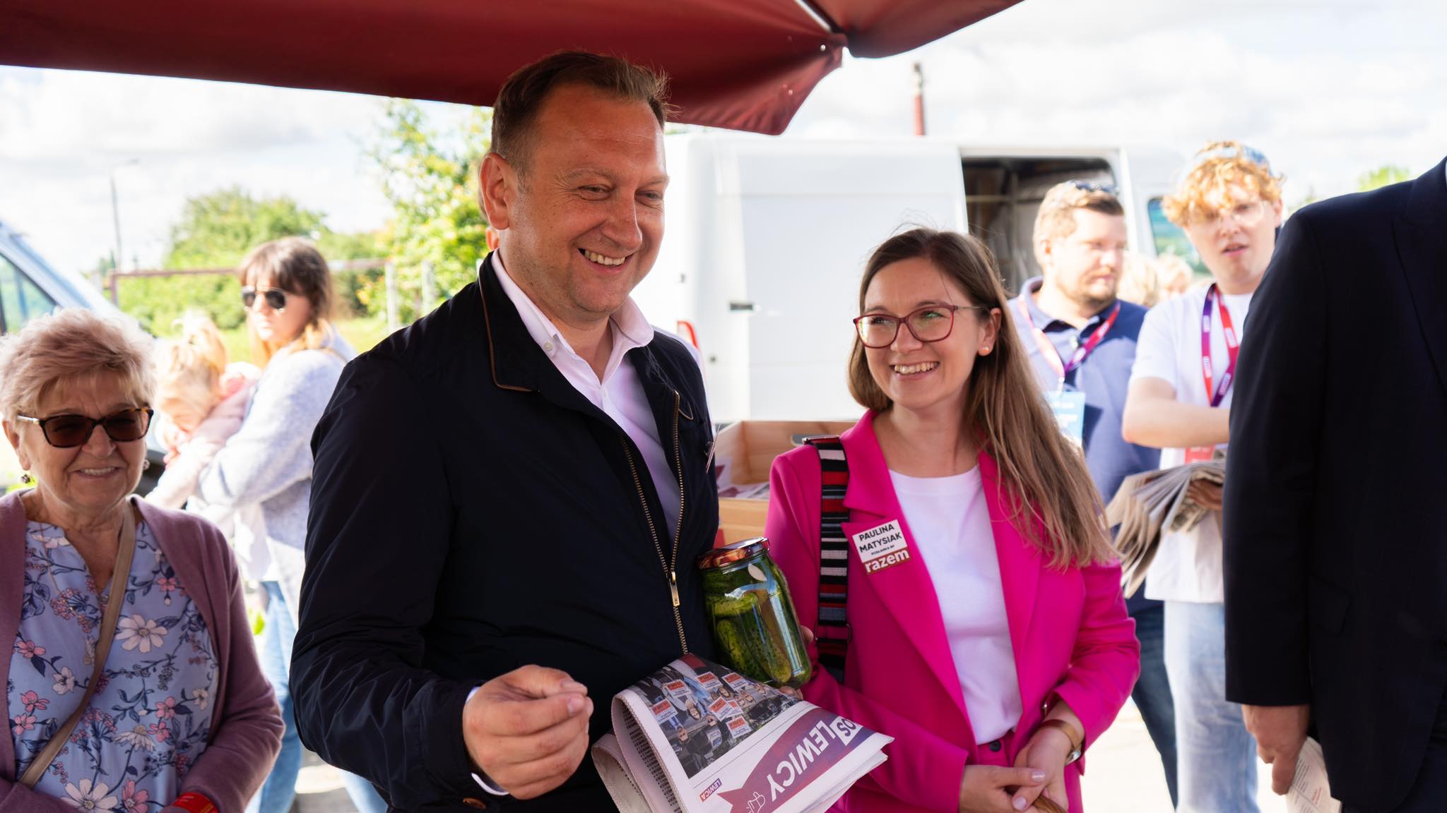 Grupa ludzi podczas wakacyjnej trasy Lewicy, uśmiechnięty mężczyzna w ciemnej kurtce trzyma gazetę i słoik, a obok niego stoi kobieta w różowym żakiecie z plakietką.