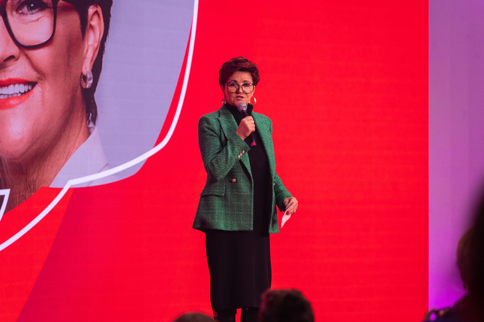 Na zdjęciu widoczna jest kobieta w zielonym żakiecie, trzymająca mikrofon i występująca na scenie przed dużym, czerwonym tłem.