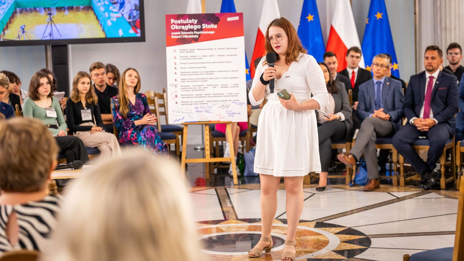 Młoda kobieta w białej sukience przemawia do zgromadzonej publiczności mikrofonem podczas podsumowania Okrągłych Stołów o zdrowiu psychicznym dzieci i młodzieży w Polsce, na tle flag Polski i Unii Europejskiej.