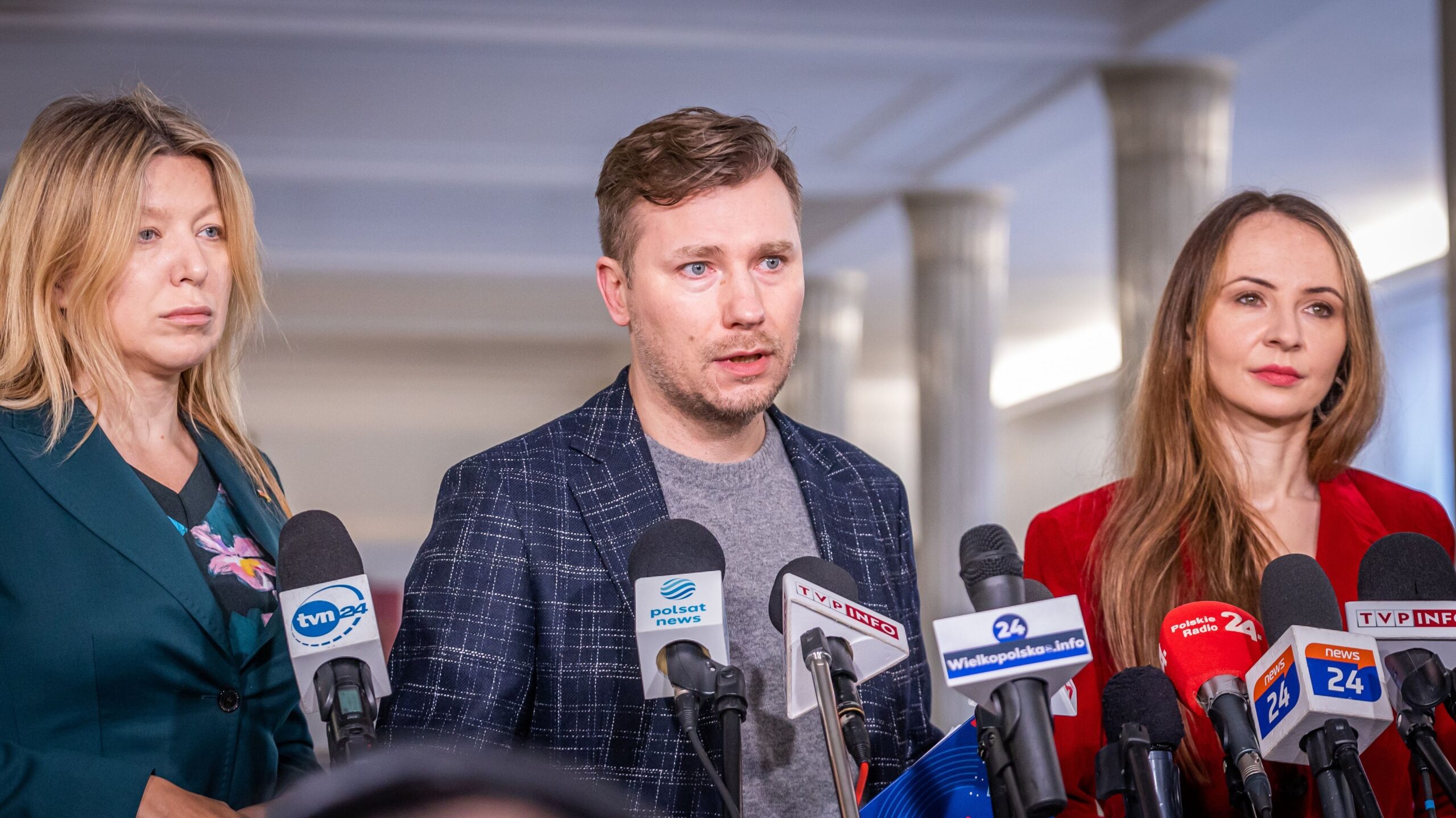 Na zdjęciu widoczni są trzy osoby uczestniczące w konferencji prasowej, stojące przed mikrofonami i wyrażające swoje stanowisko.