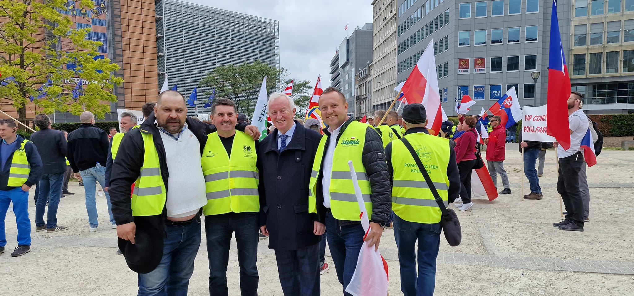 Grupa osób w żółtych kamizelkach odblaskowych stoi na placu w Brukseli podczas protestu rolników, trzymając flagi i banery, z nowoczesnymi budynkami w tle.
