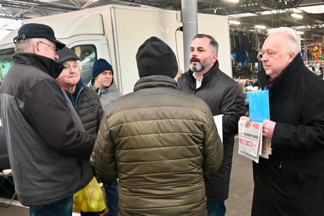 Grupa mężczyzn rozmawia na targu, przy kupionych towarach, w tle widać pojazdy i stragany.