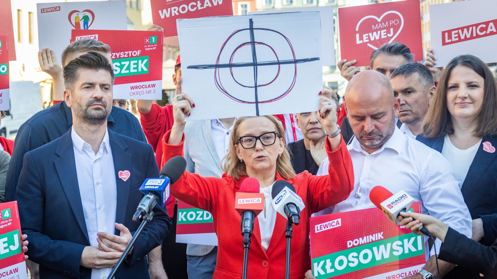 Kilkudziesięciu działaczy Lewicy podczas konferencji prasowej i demonstracji, kobieta w czerwonym żakiecie przemawia do mikrofonów, trzymając plakaty z napisem LEWICA.