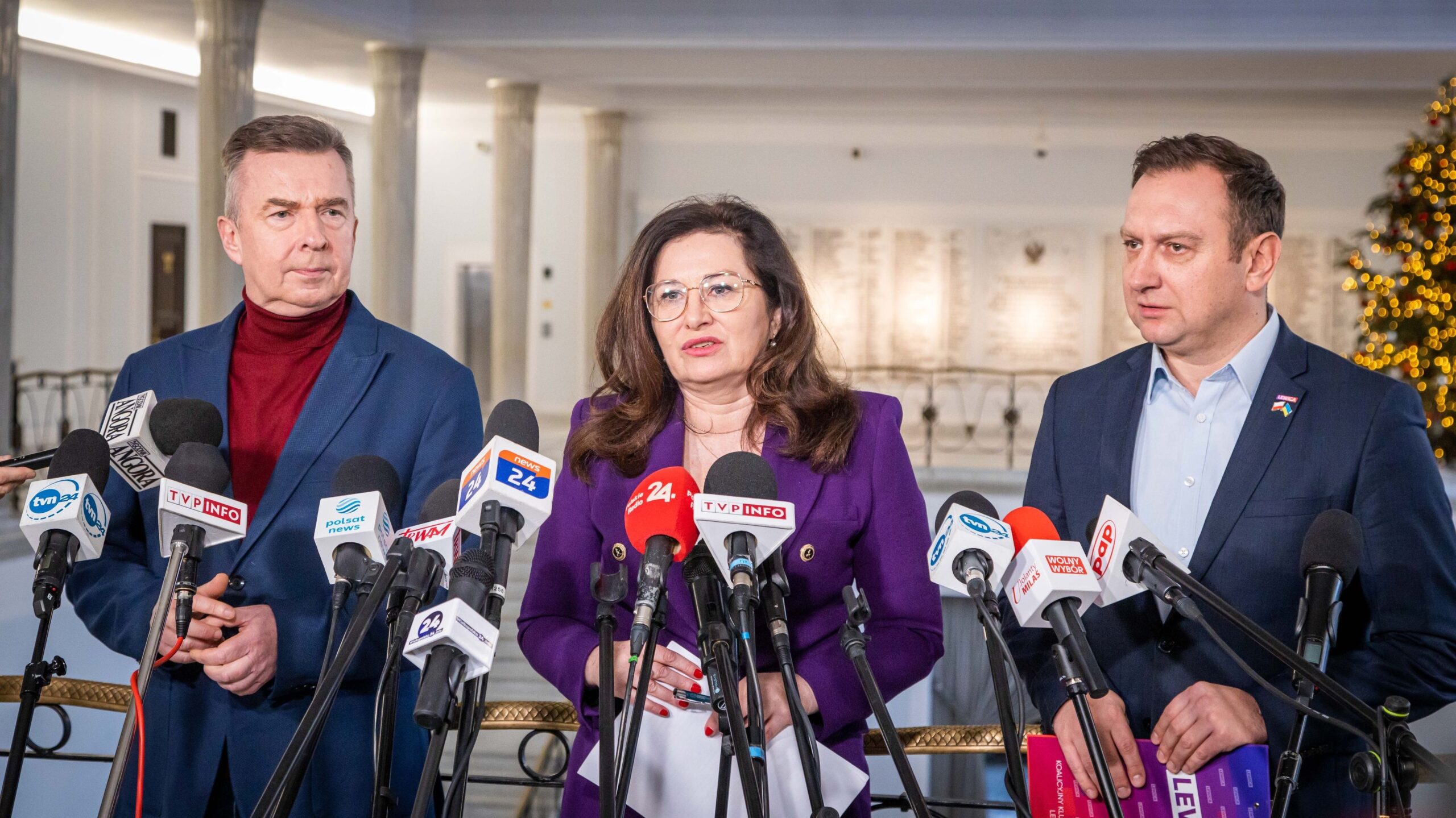 Na zdjęciu widoczni są trzej politycy podczas konferencji prasowej, z mikrofonami i kamerami dookoła, w tle ozdobione drzewko świąteczne.