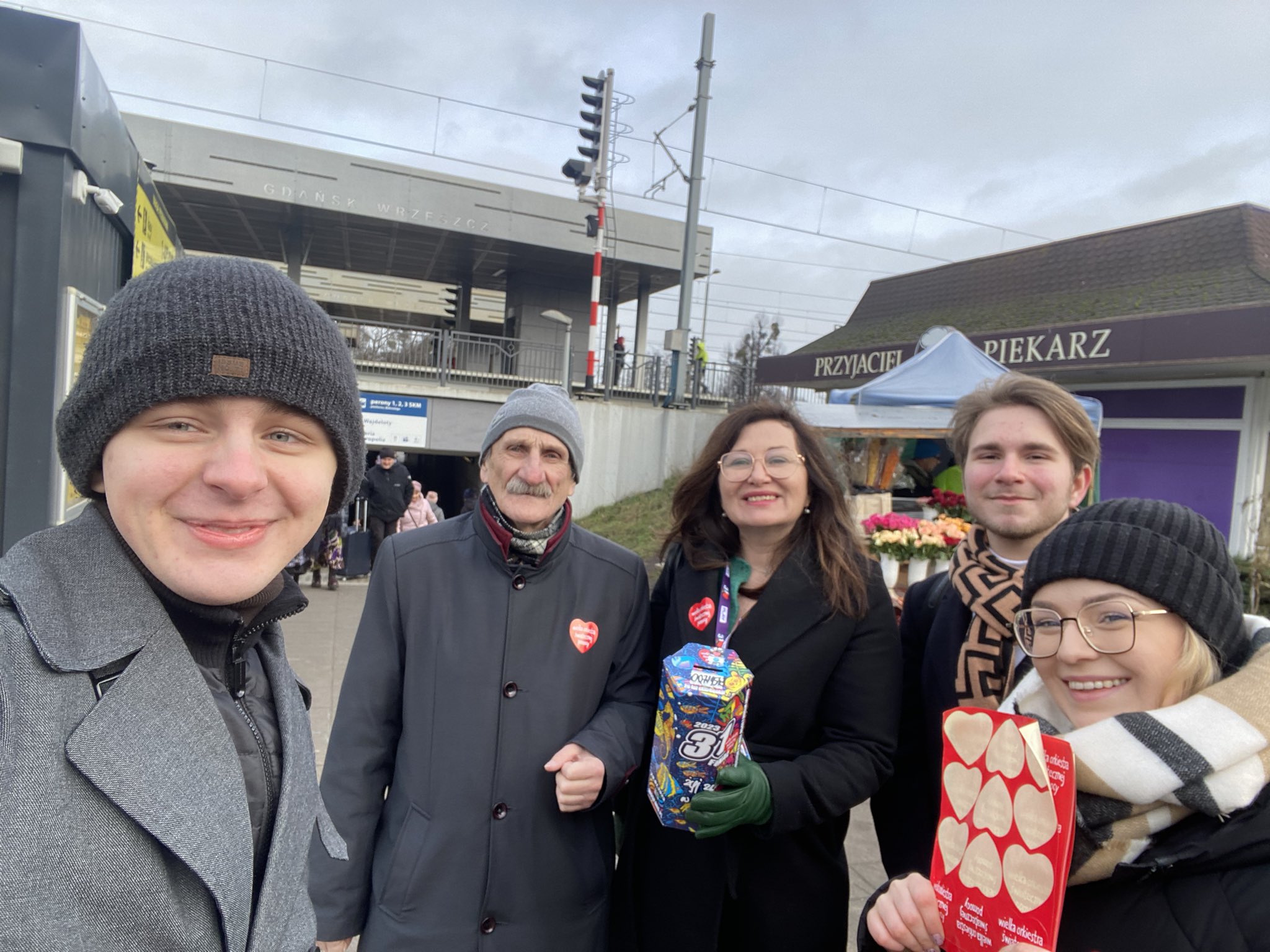 Pięć osób w zimowych ubraniach na tle stacji kolejowej podczas WOŚP 2023, uśmiechniętych do zdjęcia, trzyma serduszka i kolorowe pudełko związane ze zbiórką.