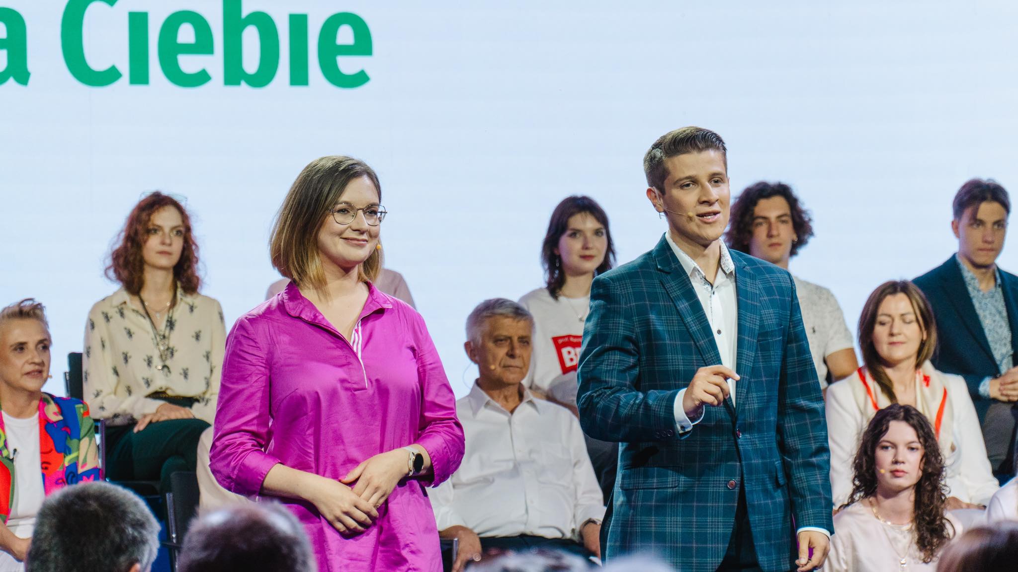 Na scenie Konwencji Europa dla Młodych grupa osób; na pierwszym planie mężczyzna w granatowym garniturze w kratę z mikrofonem oraz kobieta w różowej bluzce.