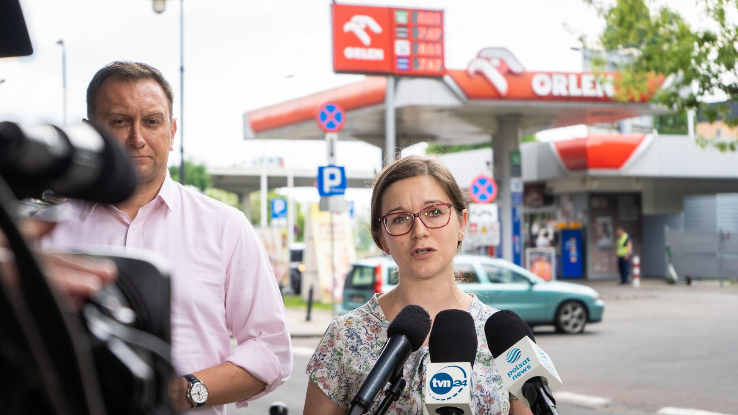 Na zdjęciu widoczna jest kobieta mówiąca do mikrofonów przed stacją benzynową Orlen, podczas gdy mężczyzna w tle obserwuje sytuację.