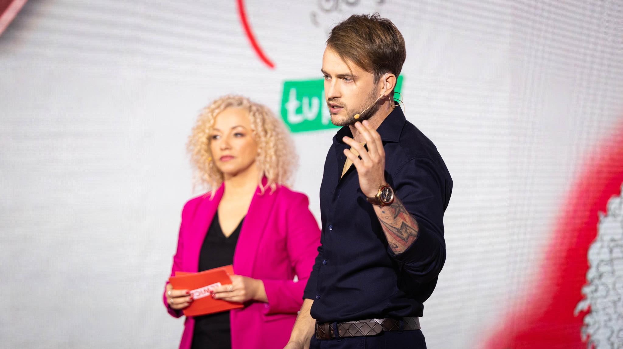 Dwóch prowadzących na scenie podczas burzy mózgów z młodzieżą; mężczyzna w granatowej koszuli z mikrofonem na uchu i tatuażem na przedramieniu przemawia, obok niego kobieta w różowym żakiecie trzyma kartki.