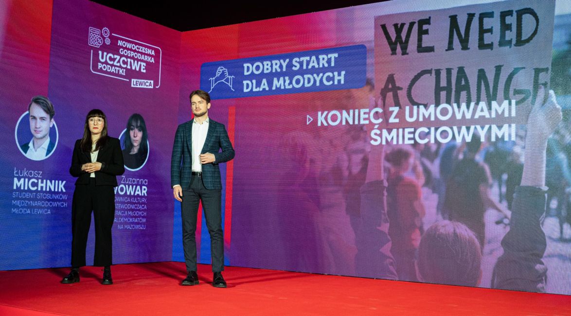 Dwóch prelegentów stoi na scenie podczas konwencji Nowej Lewicy, a w tle widać duży, kolorowy ekran z hasłami kampanii.