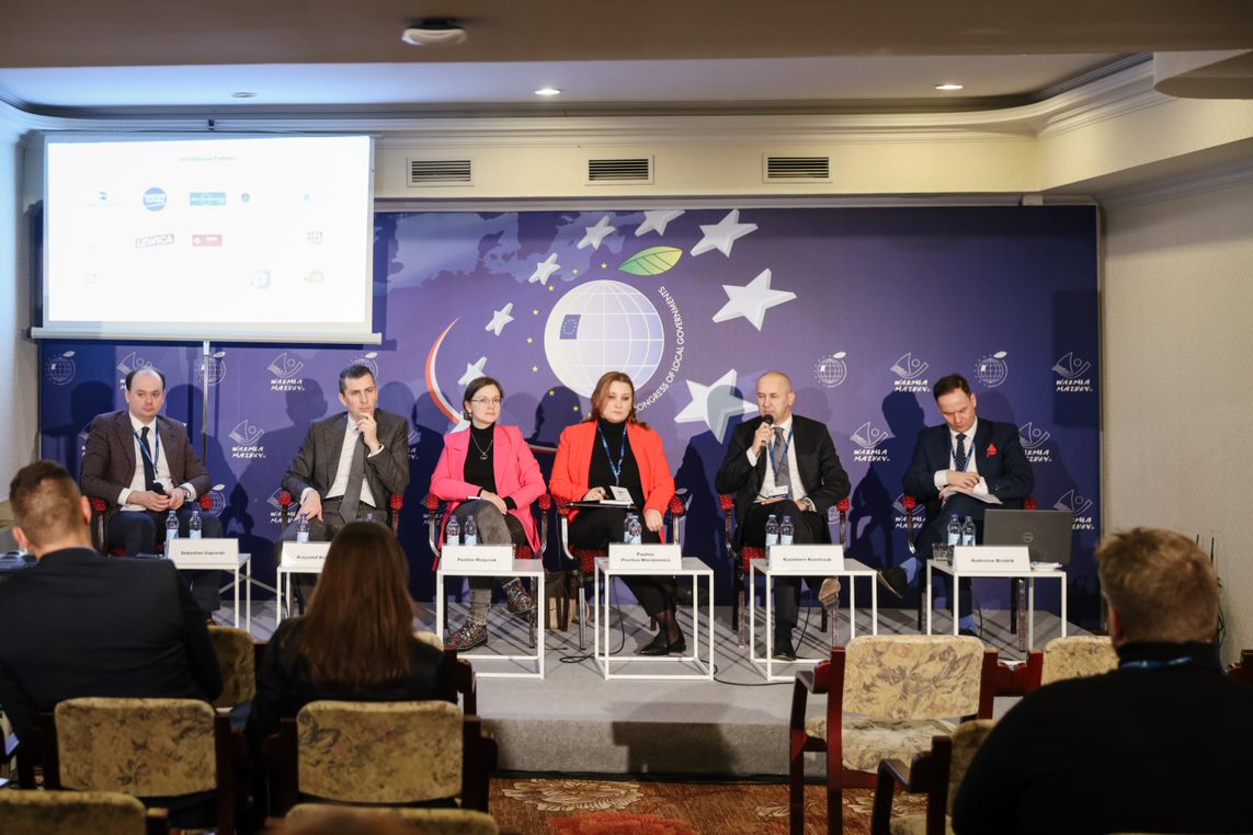 Na zdjęciu widać panel dyskusyjny z sześcioma mówcami siedzącymi przy stole w sali konferencyjnej, z logo wydarzenia w tle.