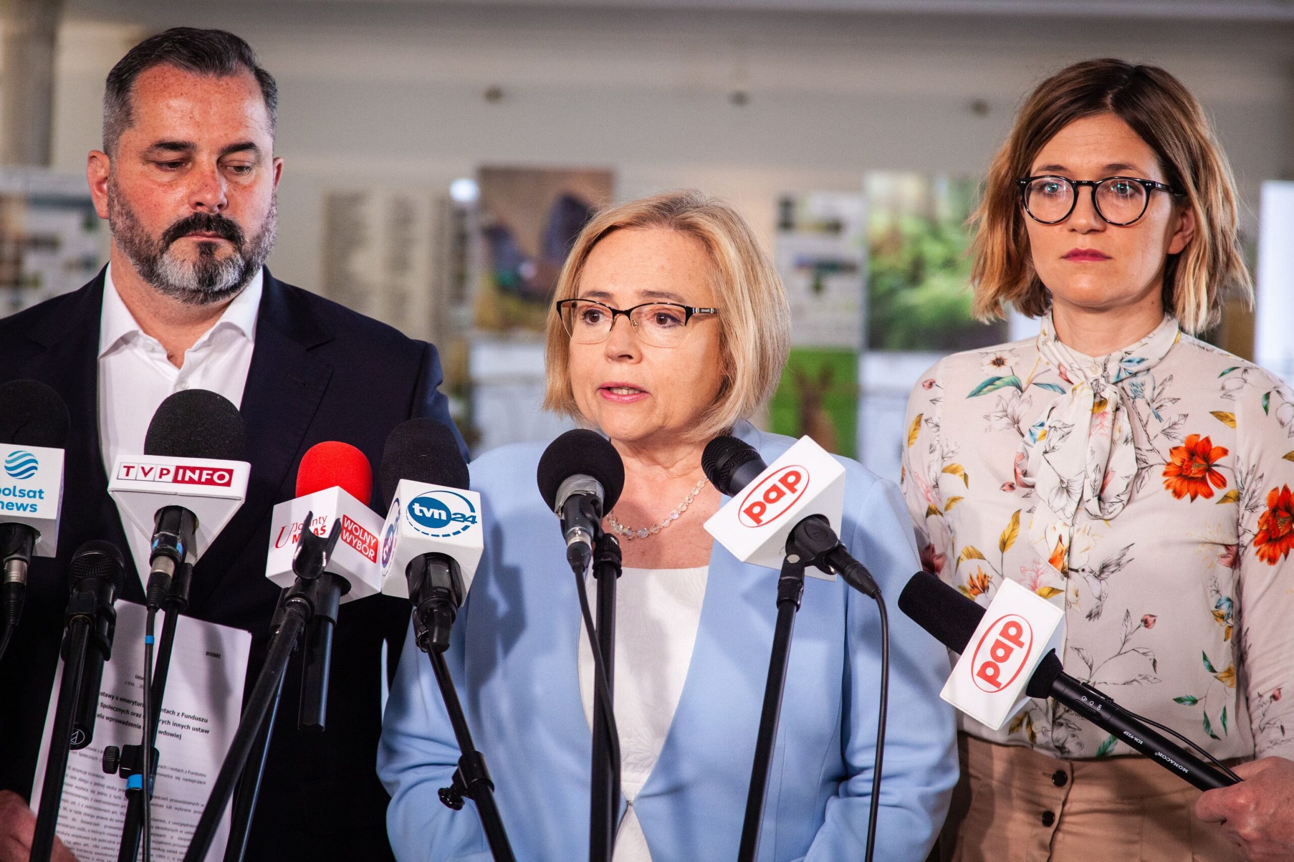 Trzy osoby stoją na konferencji prasowej z licznymi mikrofonami; pośrodku przemawia kobieta w jasnoniebieskim żakiecie, a kontekst dotyczy programu Bezpieczny Senior (druga waloryzacja emerytur, renta wdowia, leki za 5 zł).