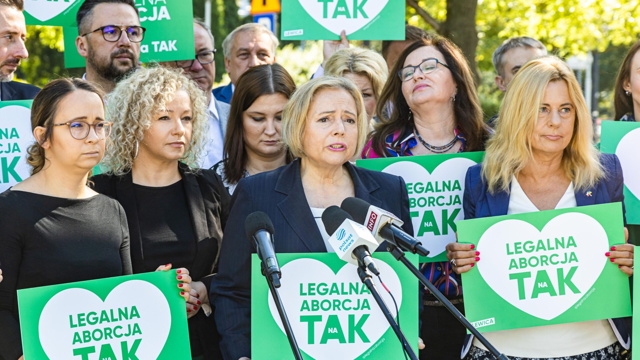 Na zdjęciu przedstawiono grupę osób, w tym kobietę mówiącą do mikrofonu, trzymających plakaty z hasłem 