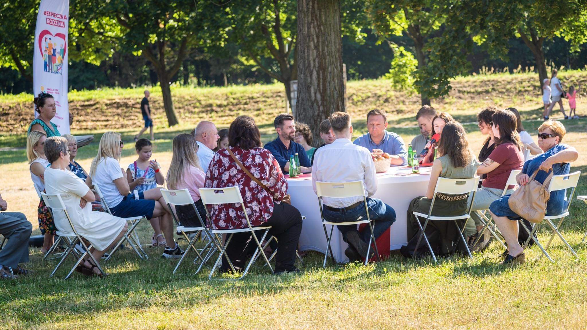 Na zdjęciu widać grupę osób siedzących przy okrągłym stole na świeżym powietrzu, uczestniczących w wydarzeniu w słoneczny dzień.