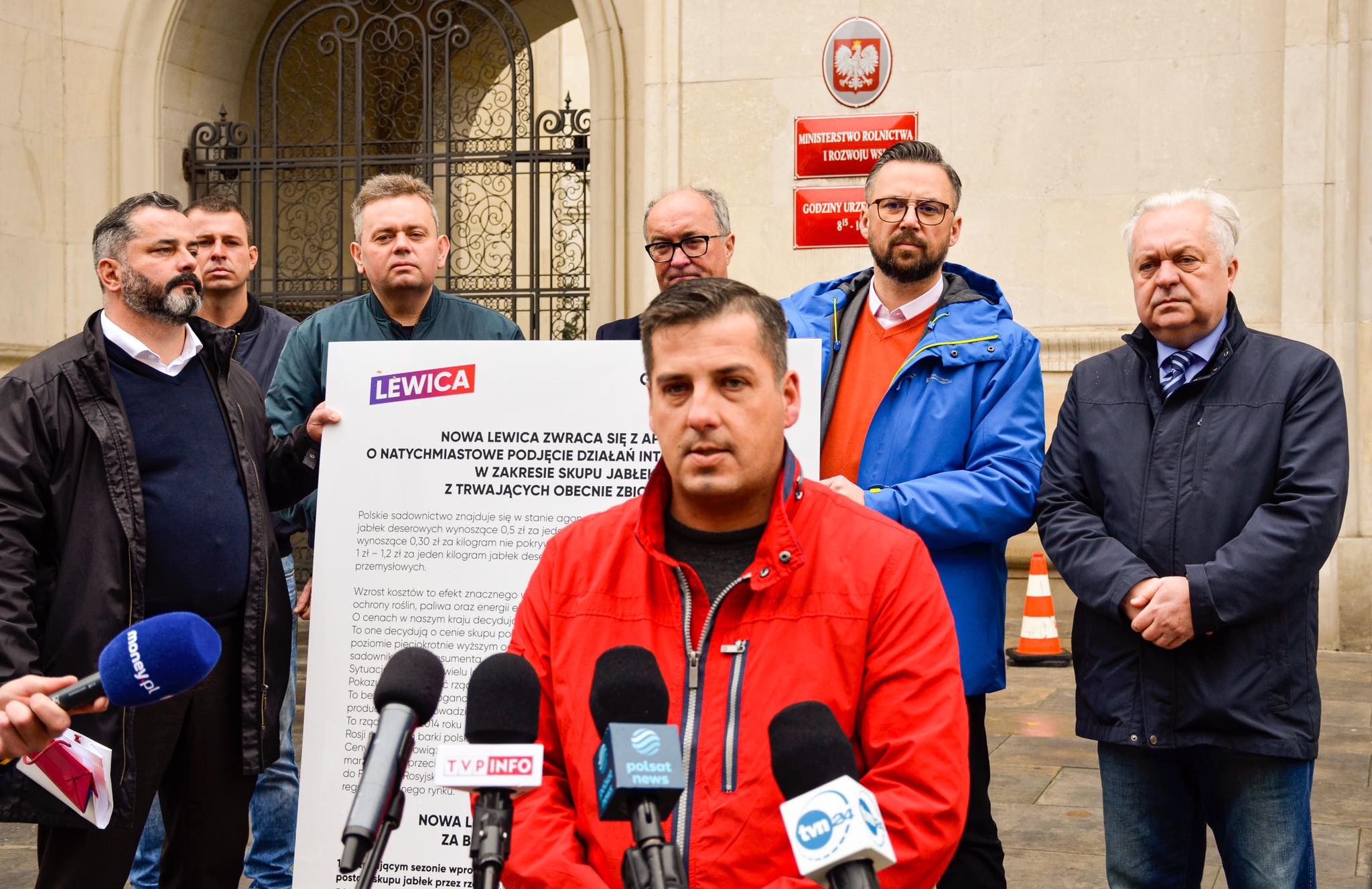 Konferencja prasowa Lewicy przed budynkiem Ministerstwa Rolnictwa i Rozwoju Wsi; na pierwszym planie mężczyzna w czerwonej kurtce przemawia do mikrofonów, za nim stoi grupa osób i duża plansza z logo Lewicy.