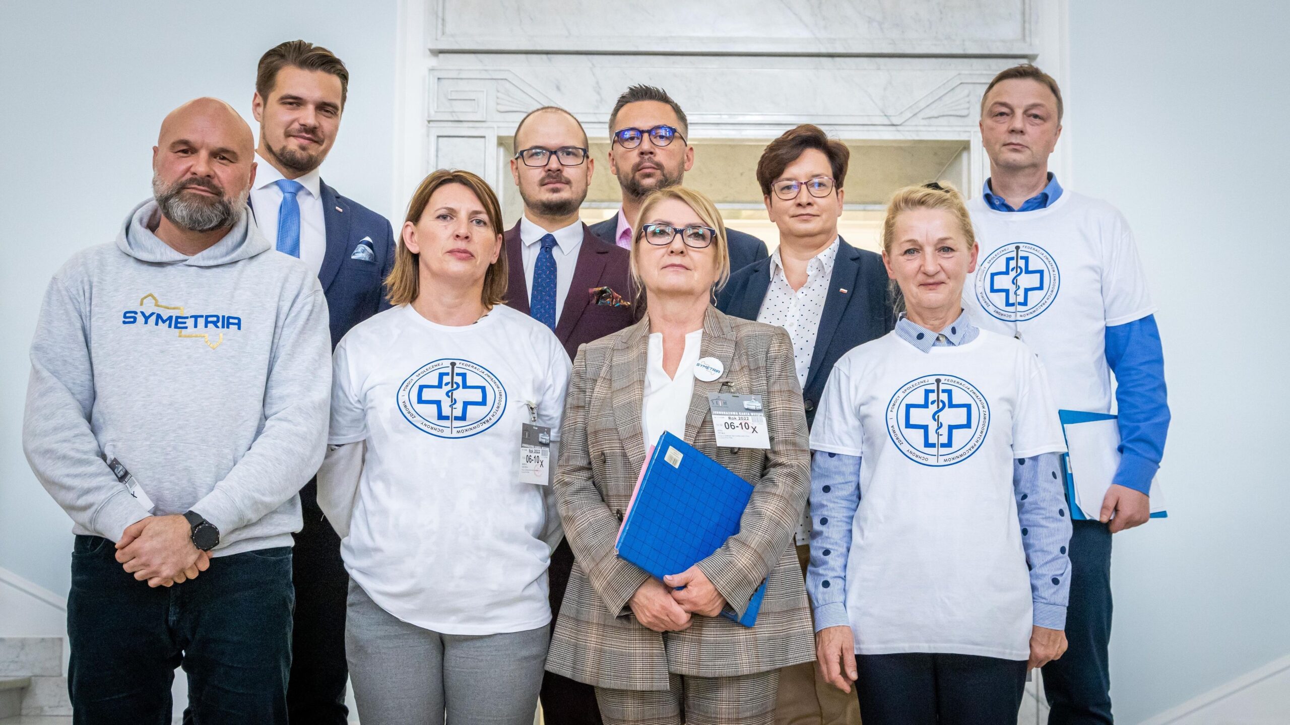 Na zdjęciu znajduje się grupa osób w różnych strojach, w tym kilka osób w białych koszulkach z niebieskim logo, stojących razem w wnętrzu budynku.