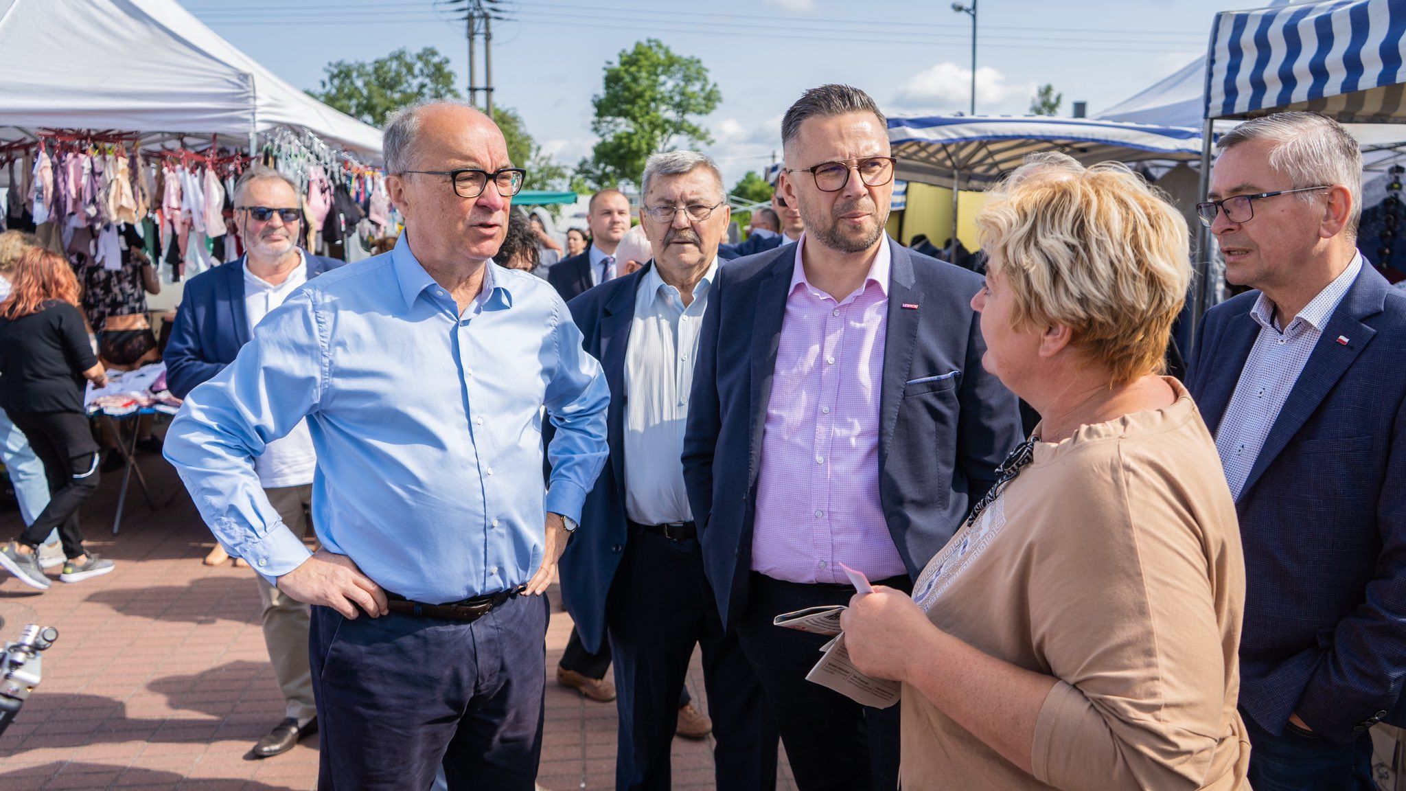 Grupa osób, w tym kilku mężczyzn w garniturach, rozmawia na zewnętrznym targu w Opolu, stojąc obok straganów z ubraniami.