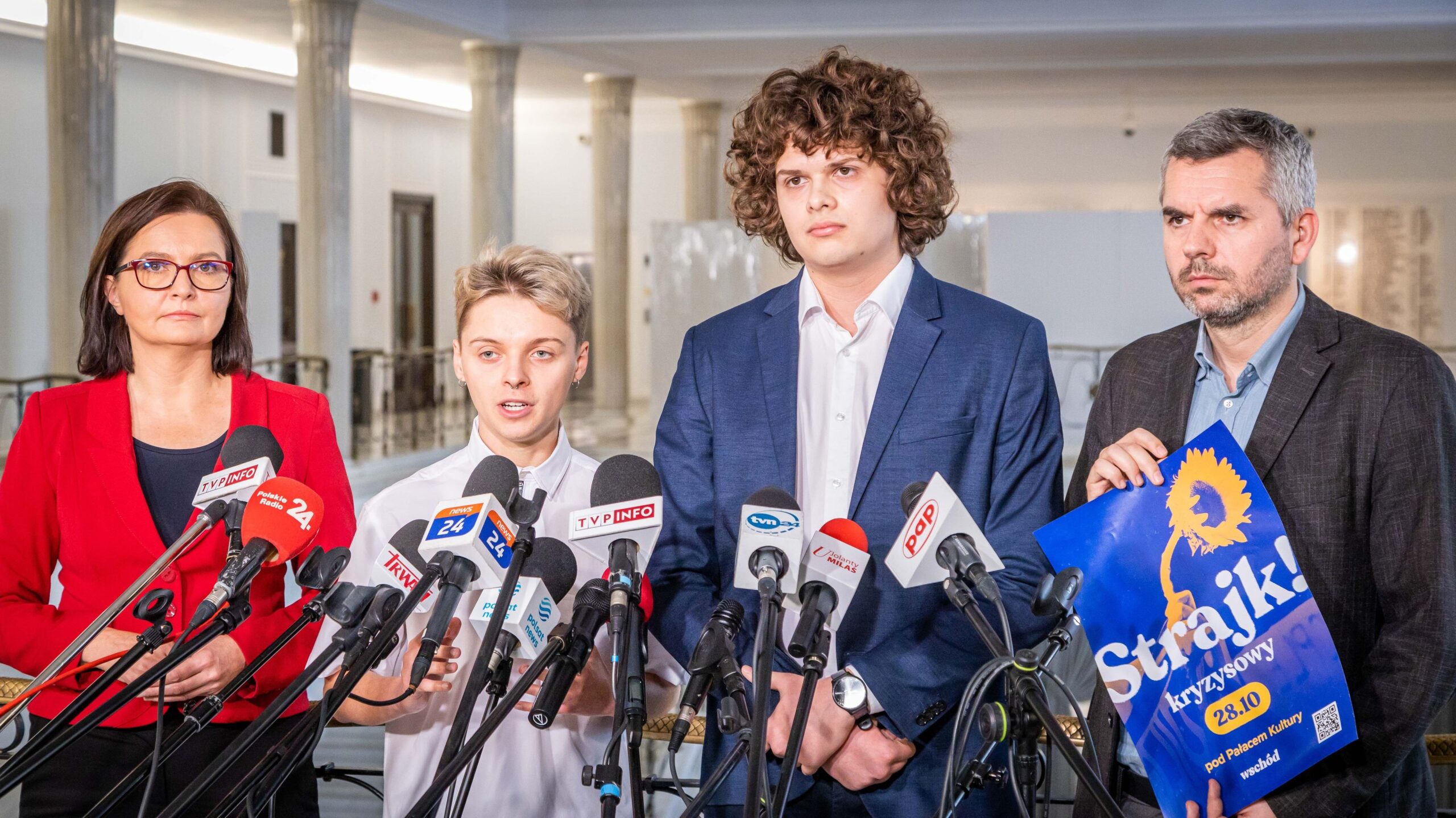 Czterech przedstawicieli różnych organizacji prowadzi konferencję prasową, trzymając plakaty i przemawiając do mediów w tle nowoczesnej architektury.