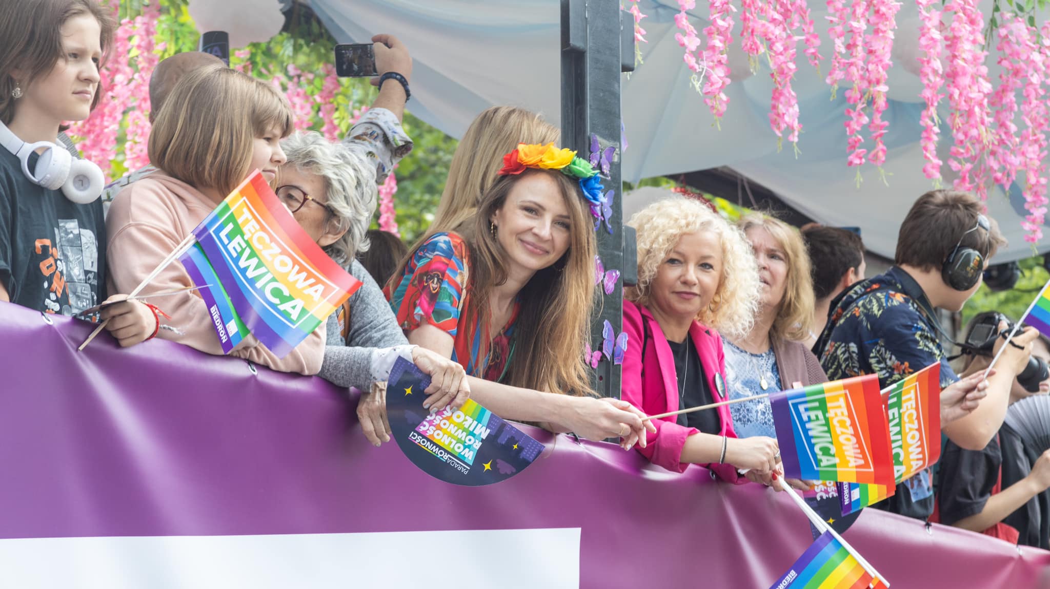 Uczestnicy Parady Równości 2023 stoją za barierką, trzymają tęczowe flagi i gadżety, a wśród nich kobieta z kwiatowym wieńcem na głowie uśmiecha się do kamery.