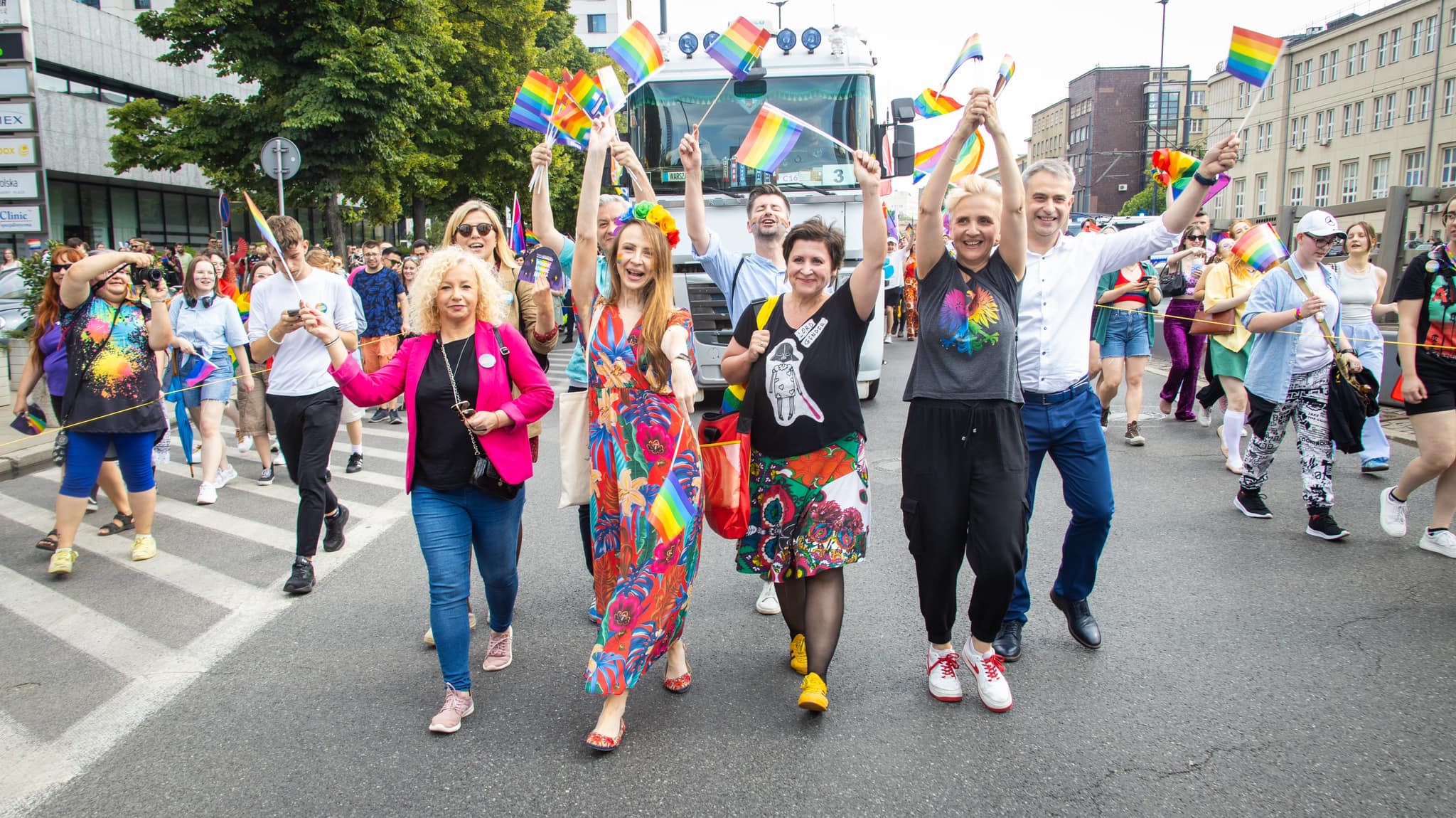 Uczestnicy Parady Równości 2023 maszerują ulicą, trzymając tęczowe flagi i uśmiechają się.