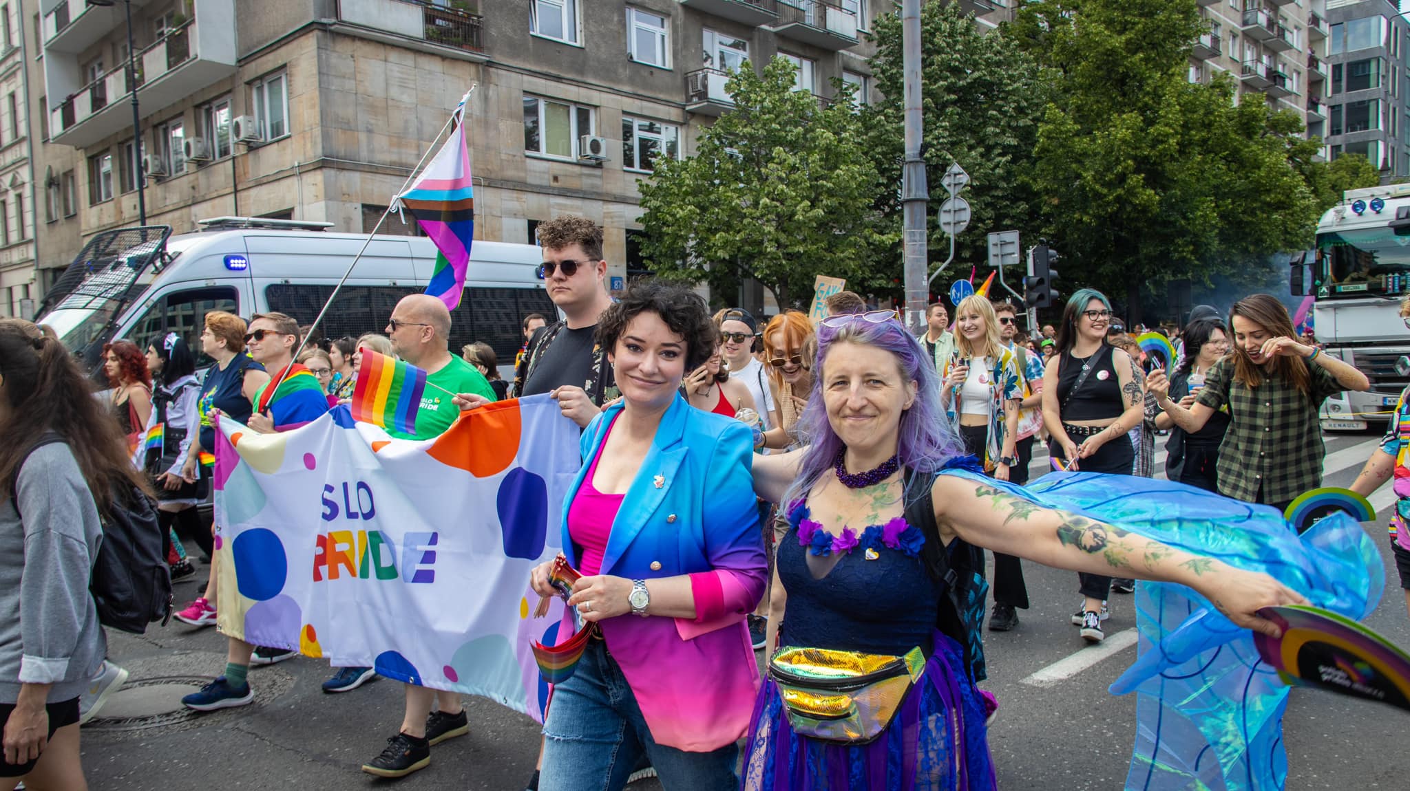 Grupa uczestników Parady Równości 2023 maszeruje ulicą, trzymając tęczowe flagi i banner z napisem SLO PRIDE.