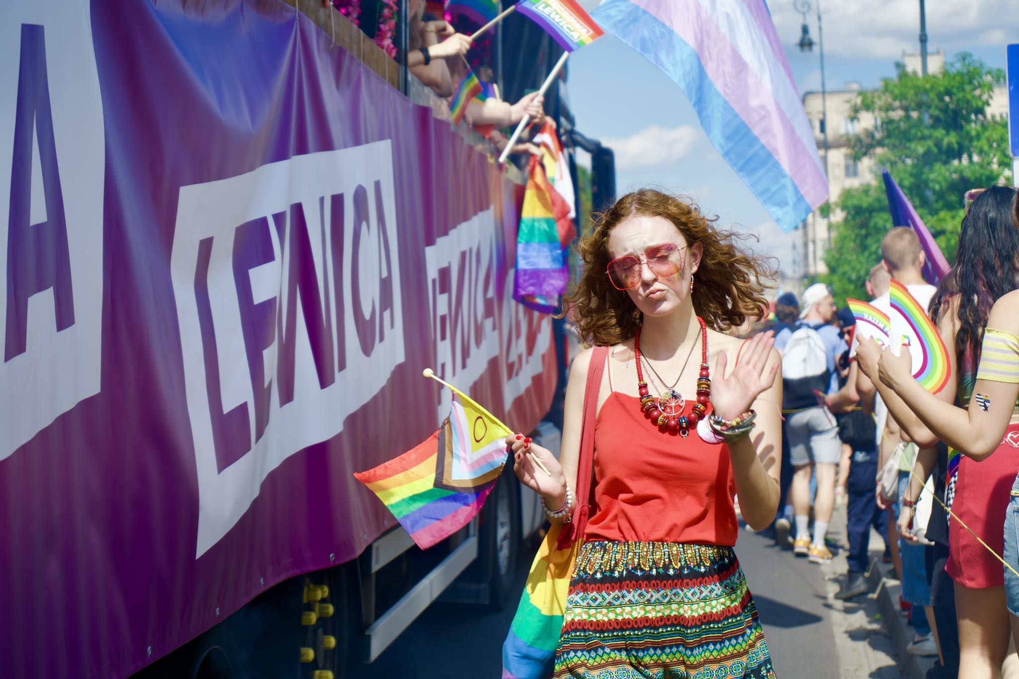 Uczestniczka Parady Równości 2024 w czerwonym topie i kolorowej spódnicy trzyma tęczową flagę i macha dłonią, stojąc obok fioletowej ciężarówki z banerem.