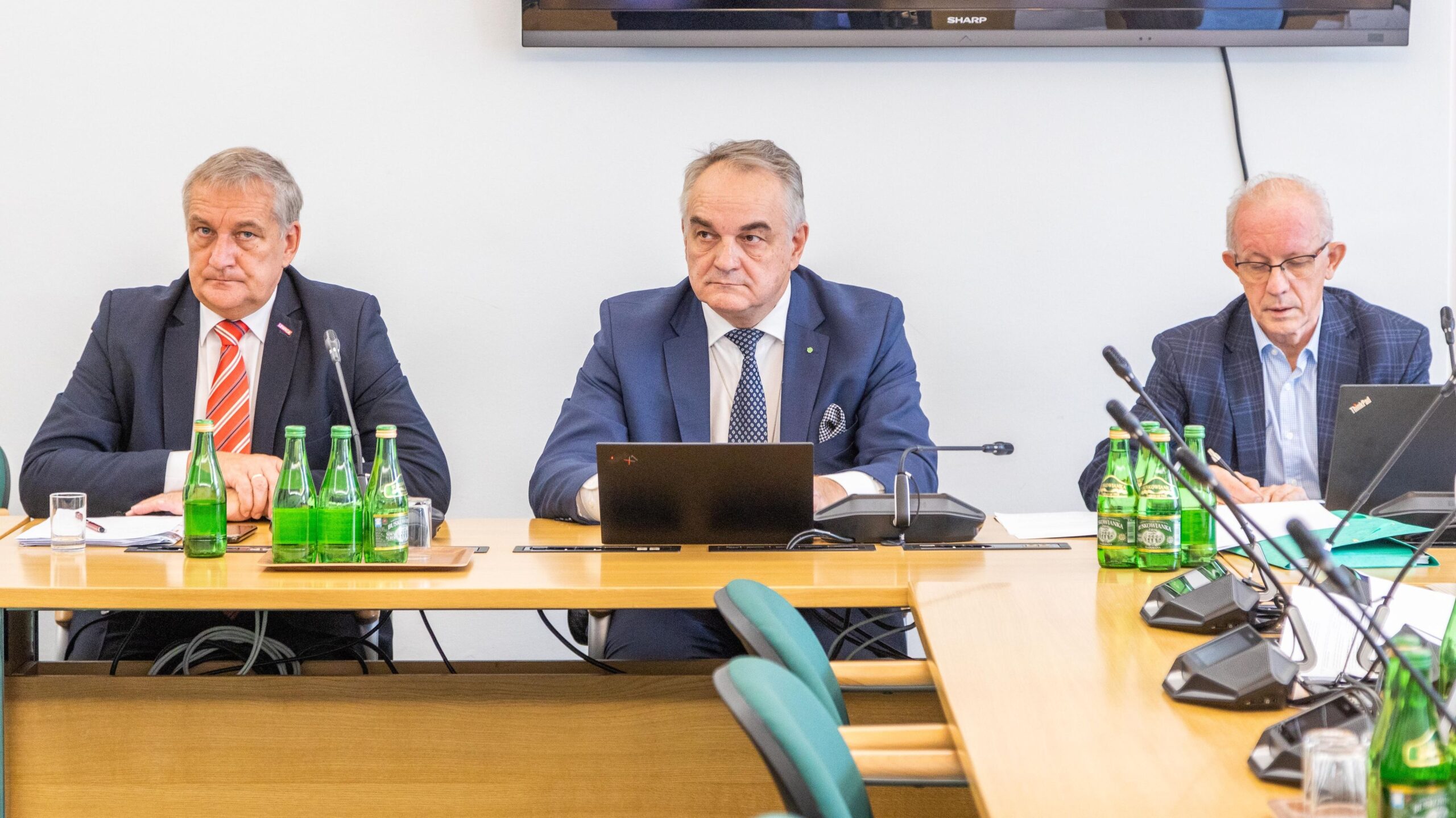 Na zdjęciu trzech mężczyzn siedzi przy stole konferencyjnym, przed nimi znajdują się butelki wody i sprzęt do nagrywania.