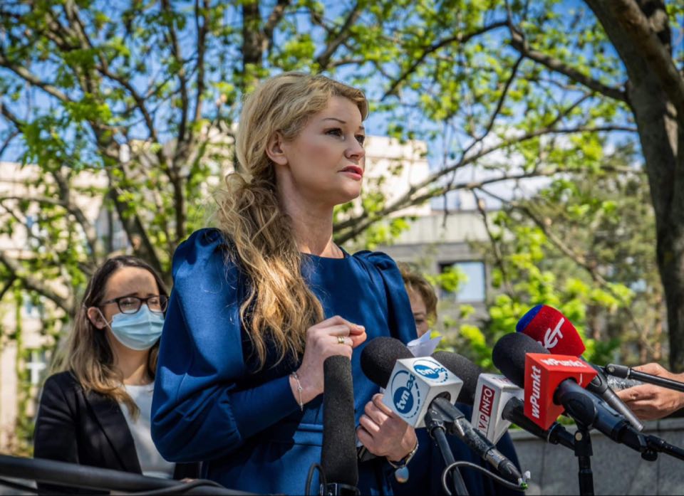 Kobieta w niebieskiej sukience przemawia podczas konferencji prasowej, stojąc przed mikrofonami dziennikarzy na tle zielonych drzew.