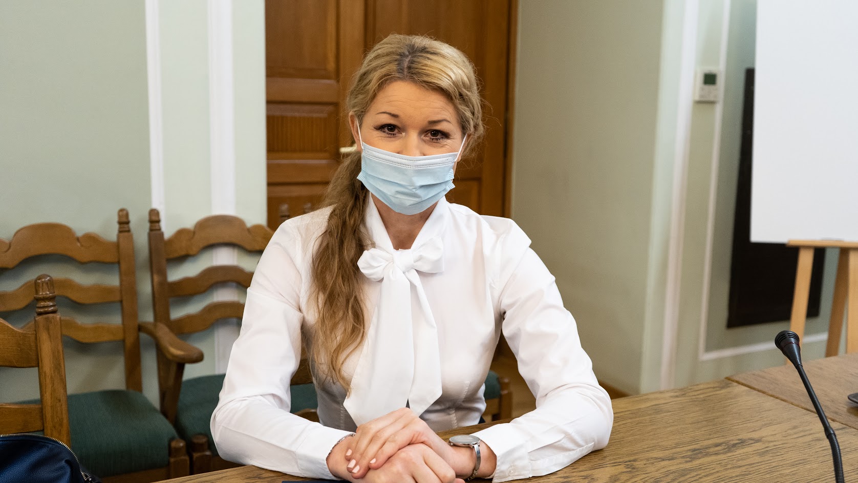Kobieta w białej bluzce z kokardą, zasłonięta maseczką, siedzi przy drewnianym stole z mikrofonem podczas spotkania, w tle widoczne krzesła i drzwi.