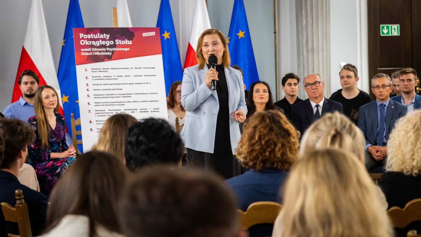 Kobieta przemawia do mikrofonu podczas konferencji, stojąc przy plakacie z napisem „Postulaty Okrągłego Stołu wokół Zdrowia Psychicznego Dzieci i Młodzieży”, w tle flagi Polski i Unii Europejskiej oraz zgromadzona publiczność.